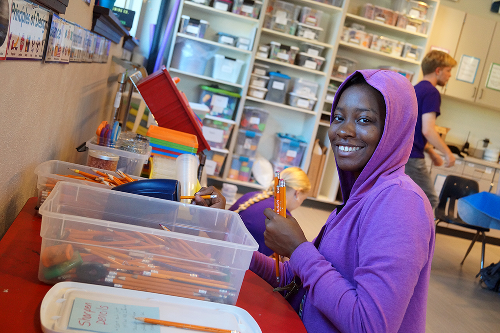 A person in a purple hoodie smiles while organizing pencils at a desk. The room is filled with educational supplies and storage containers. Another person is seen in the background, also engaged in classroom activities.