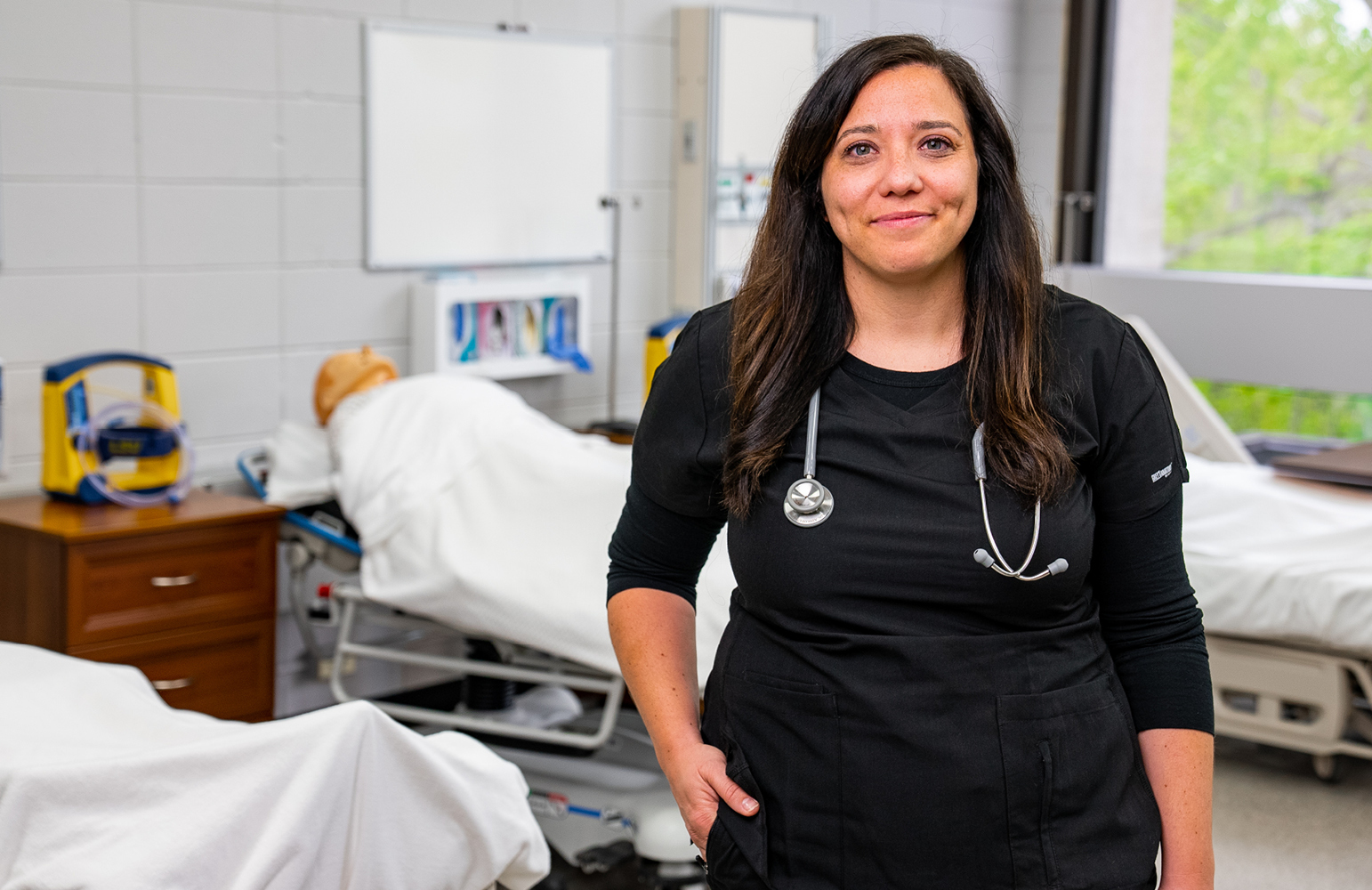 A woman wearing a black scrub top with a stethoscope around her neck stands in a medical training room. Hospital beds with mannequins are visible in the background. The room is well-lit with windows and medical equipment.