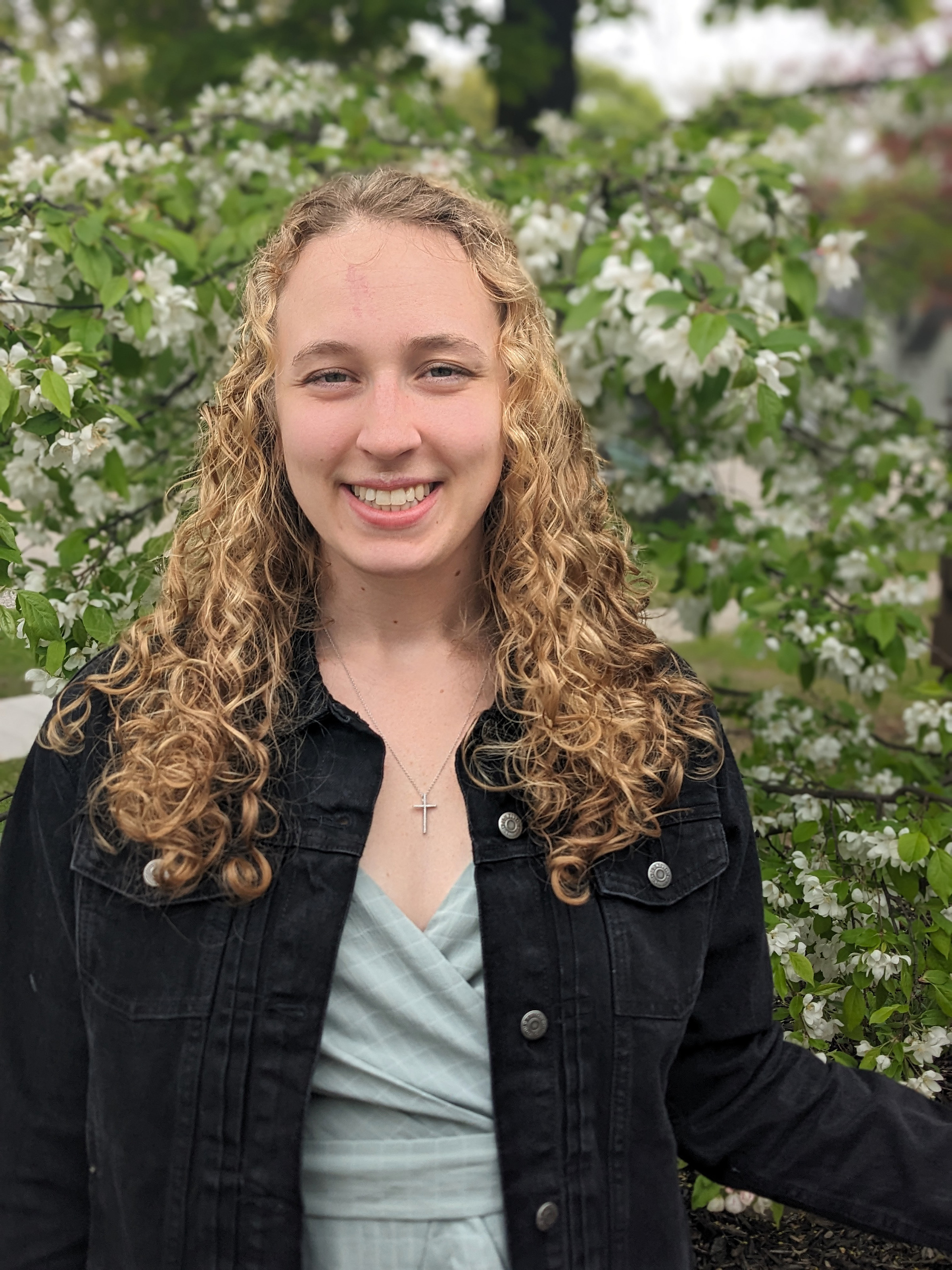 A woman with curly blonde hair, wearing a light blue top and a black jacket, is standing in front of a white flowering tree. She is smiling at the camera and wearing a cross necklace.