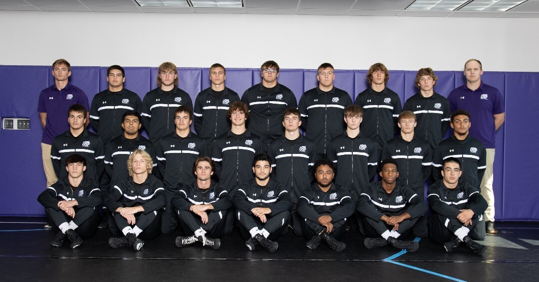 A group of young men in athletic tracksuits poses for a group photo in a gymnasium. In the back row, two men in purple polo shirts stand on either end. The team is arranged in three rows, with the front row seated cross-legged, the middle row kneeling, and the back row standing.