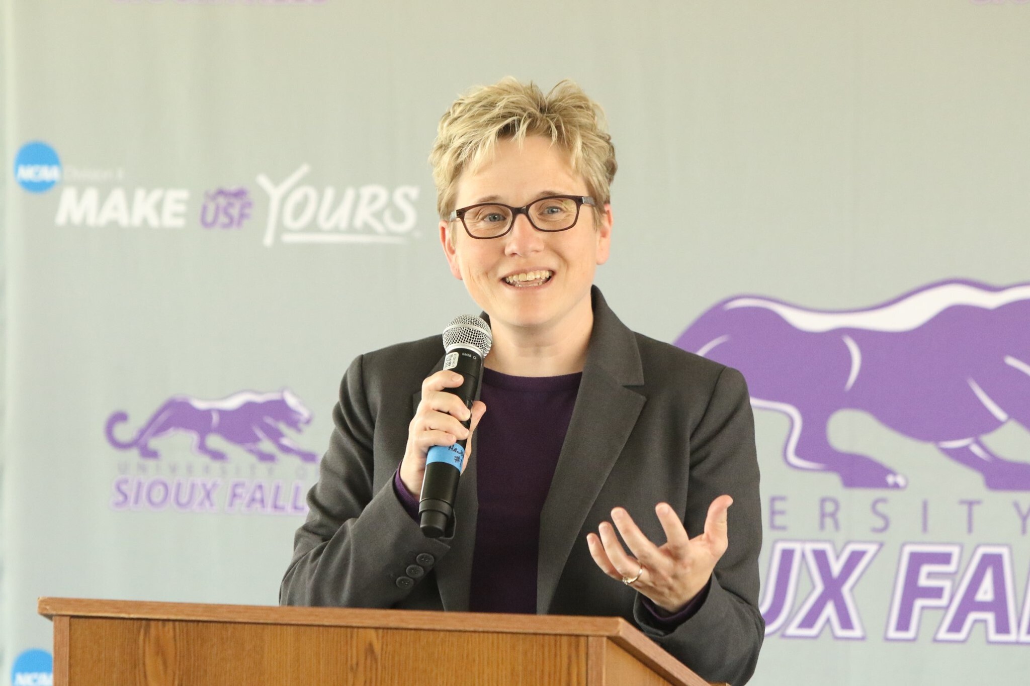 A person with short blonde hair, wearing glasses and a suit, speaks into a microphone while standing at a podium. The backdrop displays the logo and text for the University of Sioux Falls, including a purple panther and the phrase "Make It Yours.