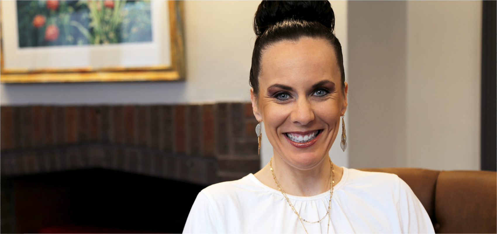 A woman with dark hair styled in a bun is smiling while sitting on a chair indoors. She is wearing a white top, long earrings, and layered necklaces. There's a framed picture and brickwork in the background.