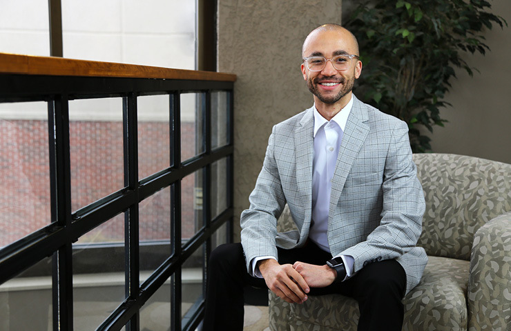 A man with glasses, wearing a gray plaid blazer and white shirt, sits on a patterned armchair by a large window. He is smiling and has his hands resting on his lap. There is a plant in the background and a wooden railing by the window.