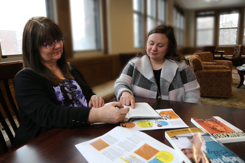 Two women sit at a table in a bright room with large windows. They are conversing and looking at a book one of them is holding open. Various documents are spread out on the table, including magazines and papers with colorful graphics.