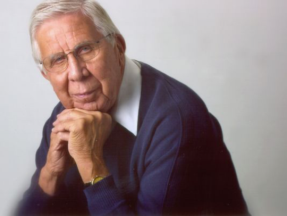 An elderly man with white hair and glasses is seated, resting his chin on his hands. He is wearing a dark blue sweater over a white collared shirt. The background is plain and light gray. His facial expression is calm and reflective.