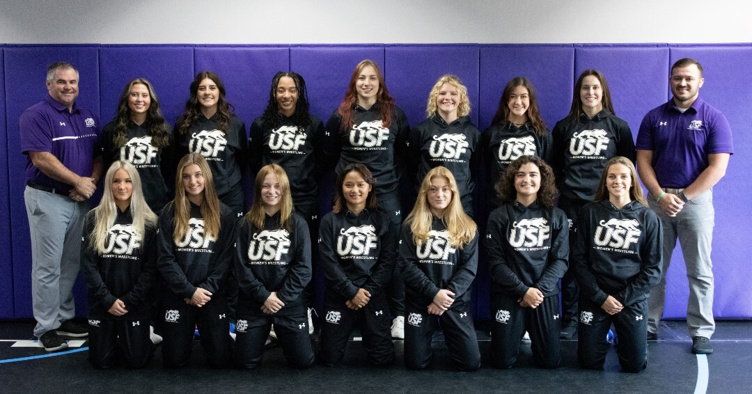 A group of fourteen women in matching black athletic attire with "USF" logo pose in front of a purple wall. They are flanked by two men in purple shirts. Some are standing while others are kneeling, all smiling at the camera.
