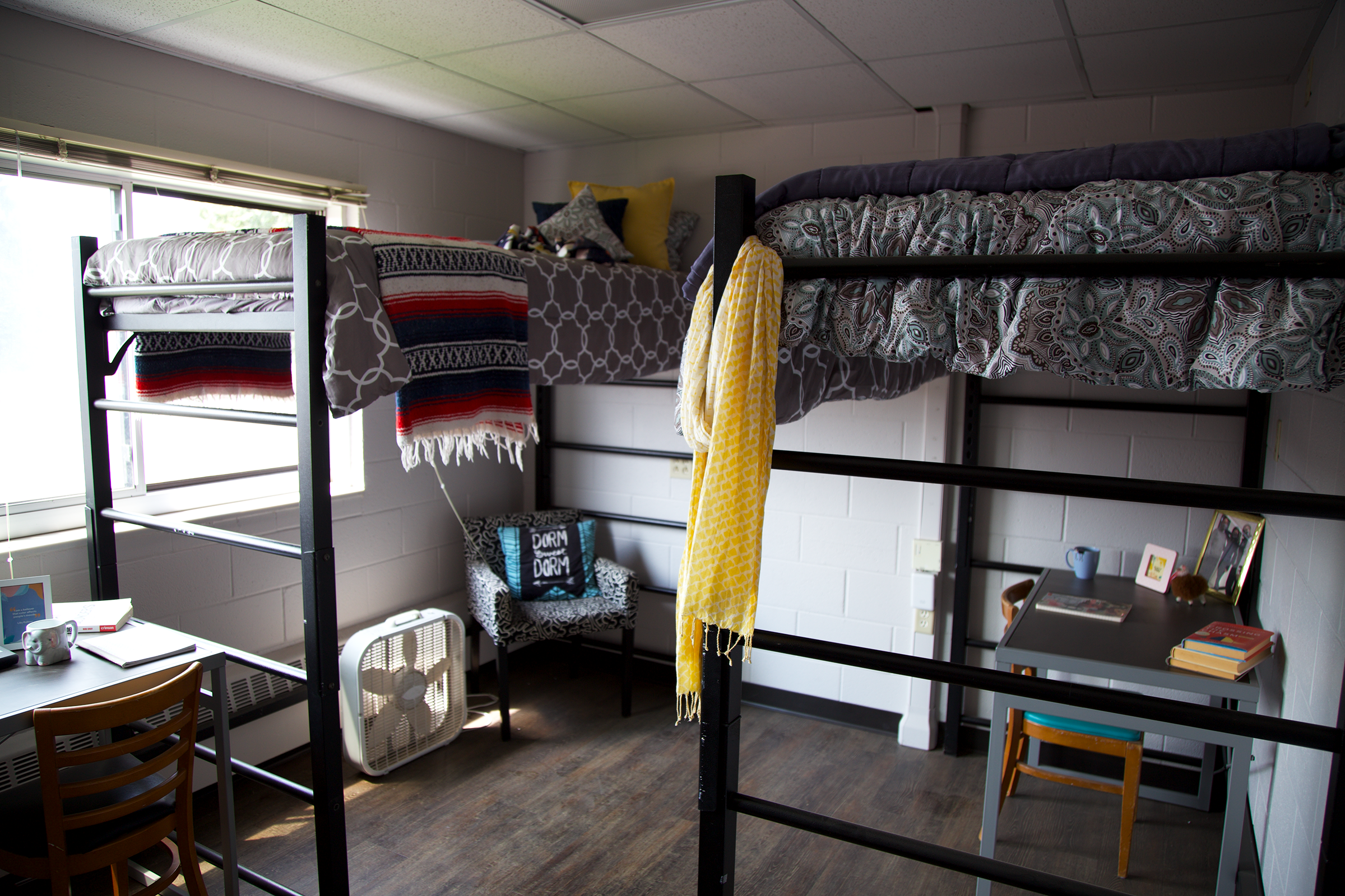 A dorm room with two lofted beds, each topped with patterned bedding. One bed has a throw blanket draped over the side. A fan and decorative pillows are beneath the beds, alongside a desk with books, a lamp, and a chair by a window.