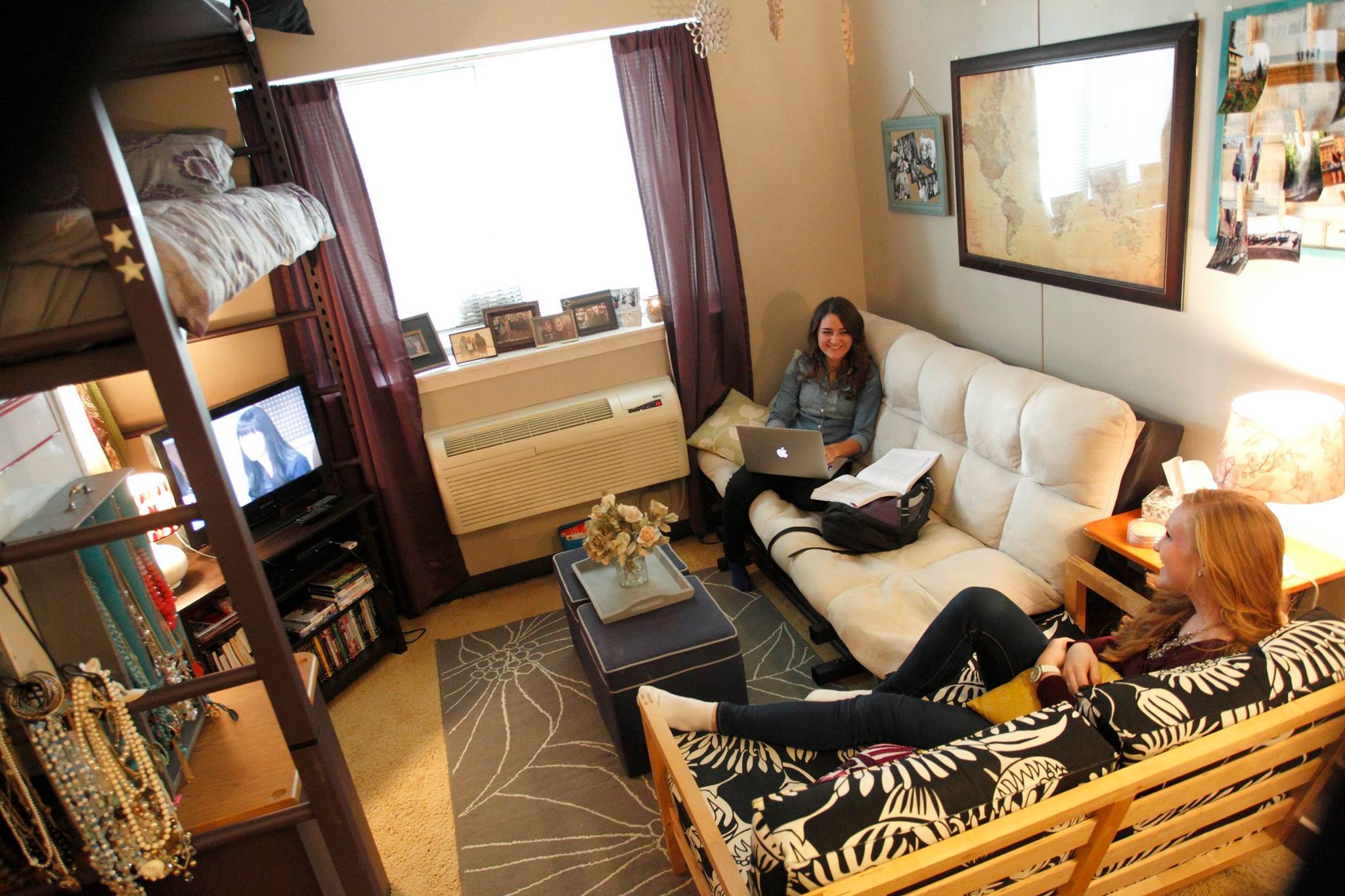 A cozy dorm room with two young women; one sits on a beige couch with a laptop, the other reclines on a black and white patterned futon. The room has a lofted bed, wall decorations, a small TV, shelves, and a window with curtains. Textbooks and frames are visible.