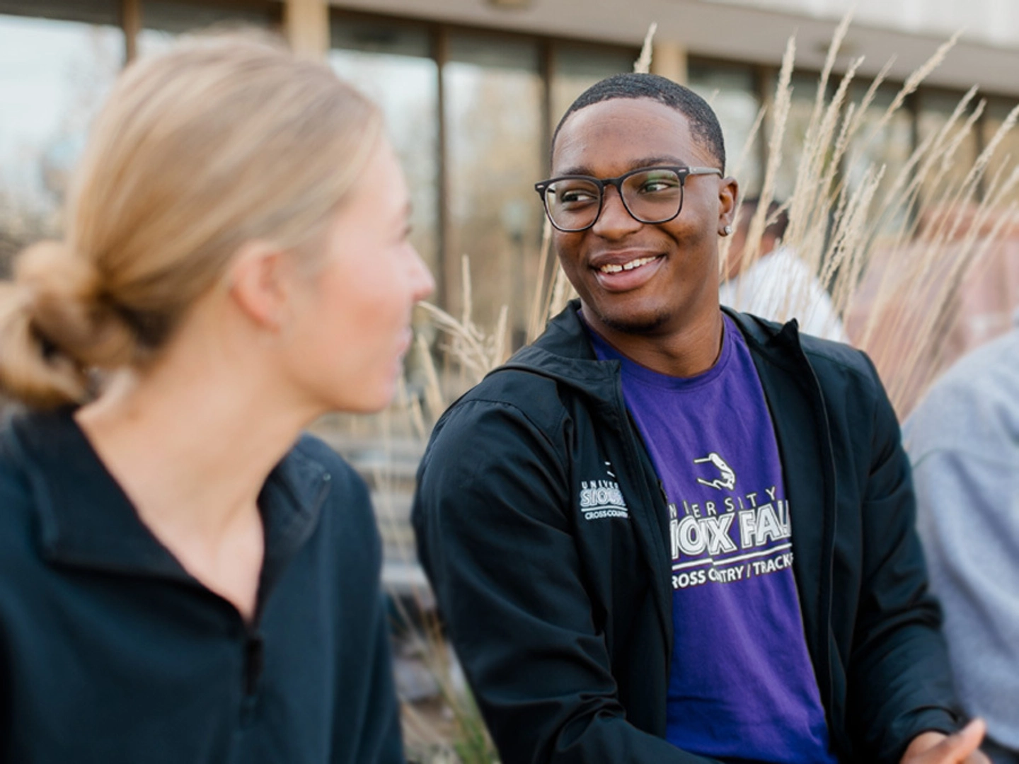 Two students smile and converse while seated in front of a campus building