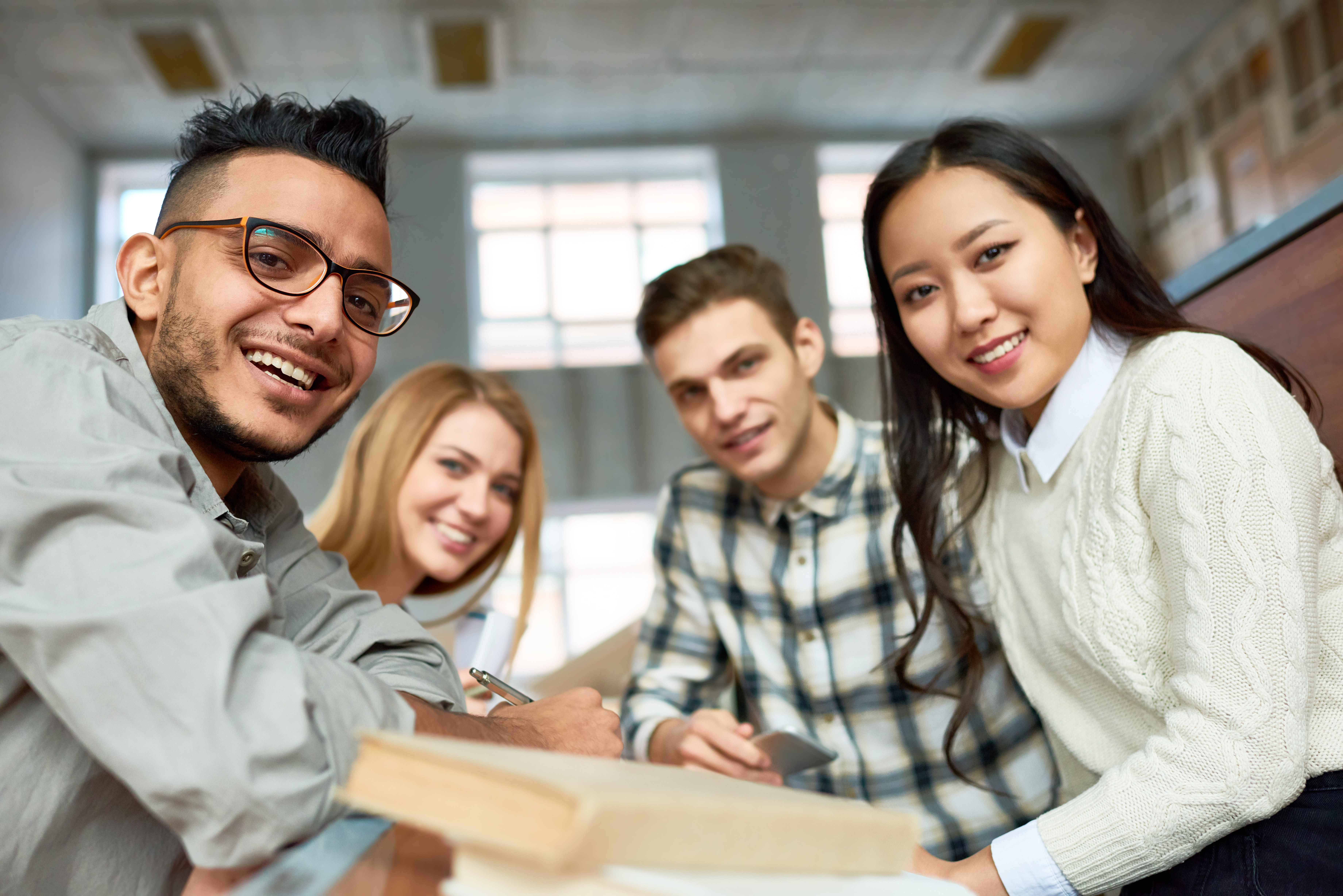 A group of four young adults, three men and one woman, sit together at a table in a bright room. They are smiling and looking at the camera. Piles of books and notebooks are on the table, suggesting a study session or collaborative work environment.