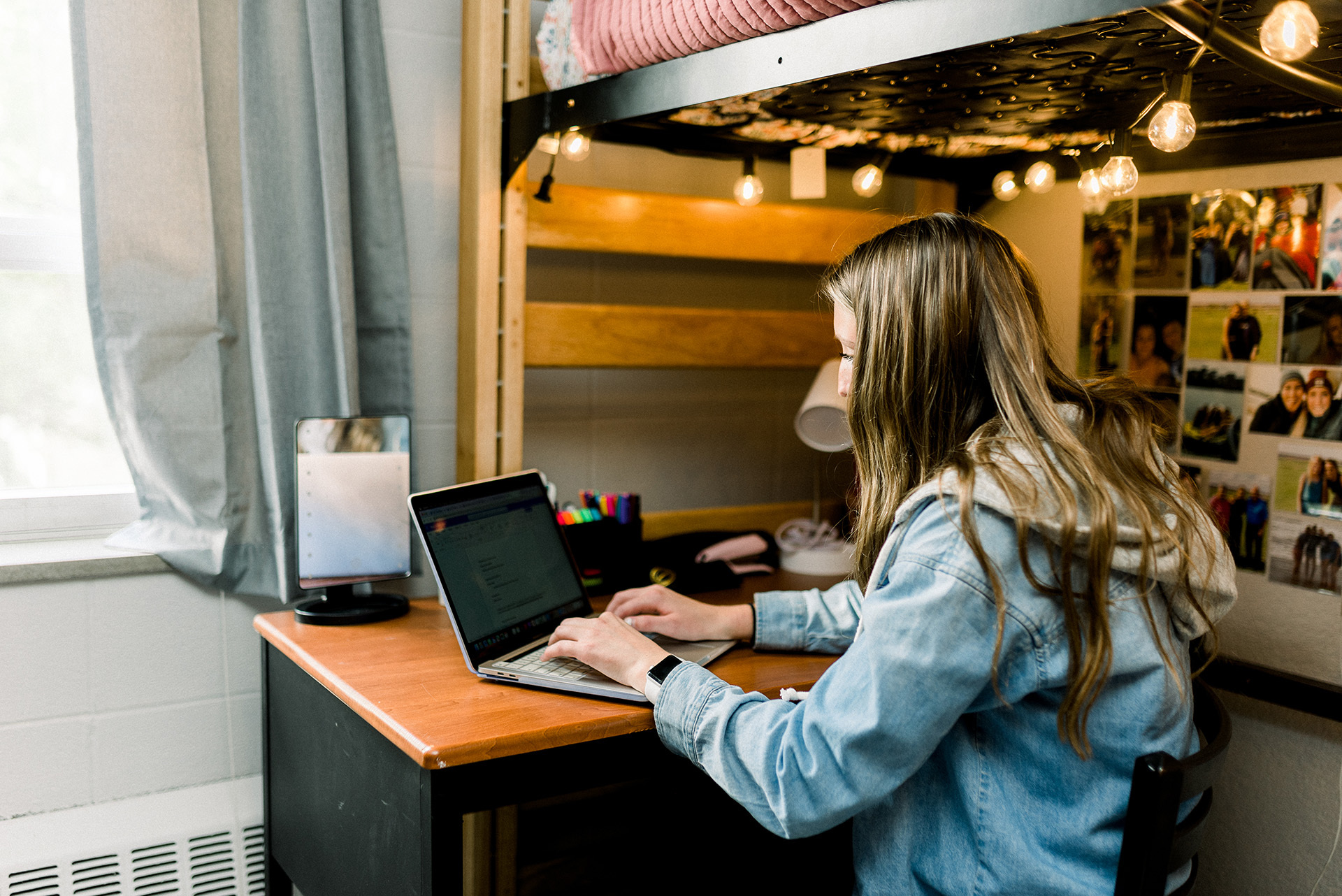 A person sits at a desk under a lofted bed, working on a laptop. The desk is decorated with string lights, photos, and stationery. Sunlight streams in through a nearby window, creating a cozy and focused atmosphere in the room.