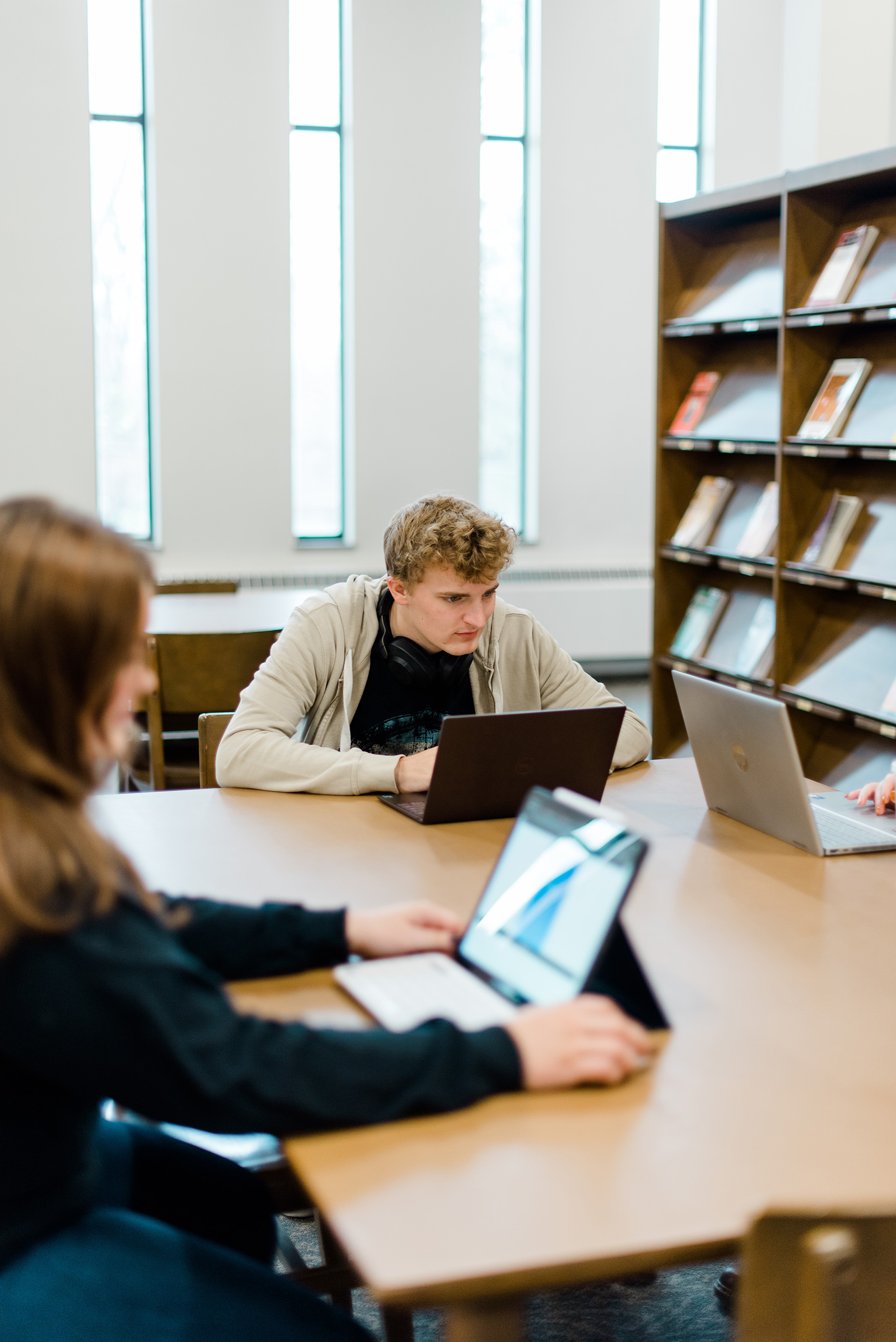 Three people are sitting at a table in a library, each working on laptops. Bookshelves with books are visible in the background, along with tall windows letting in daylight. The focus is on a person in the middle, looking intently at their laptop screen.