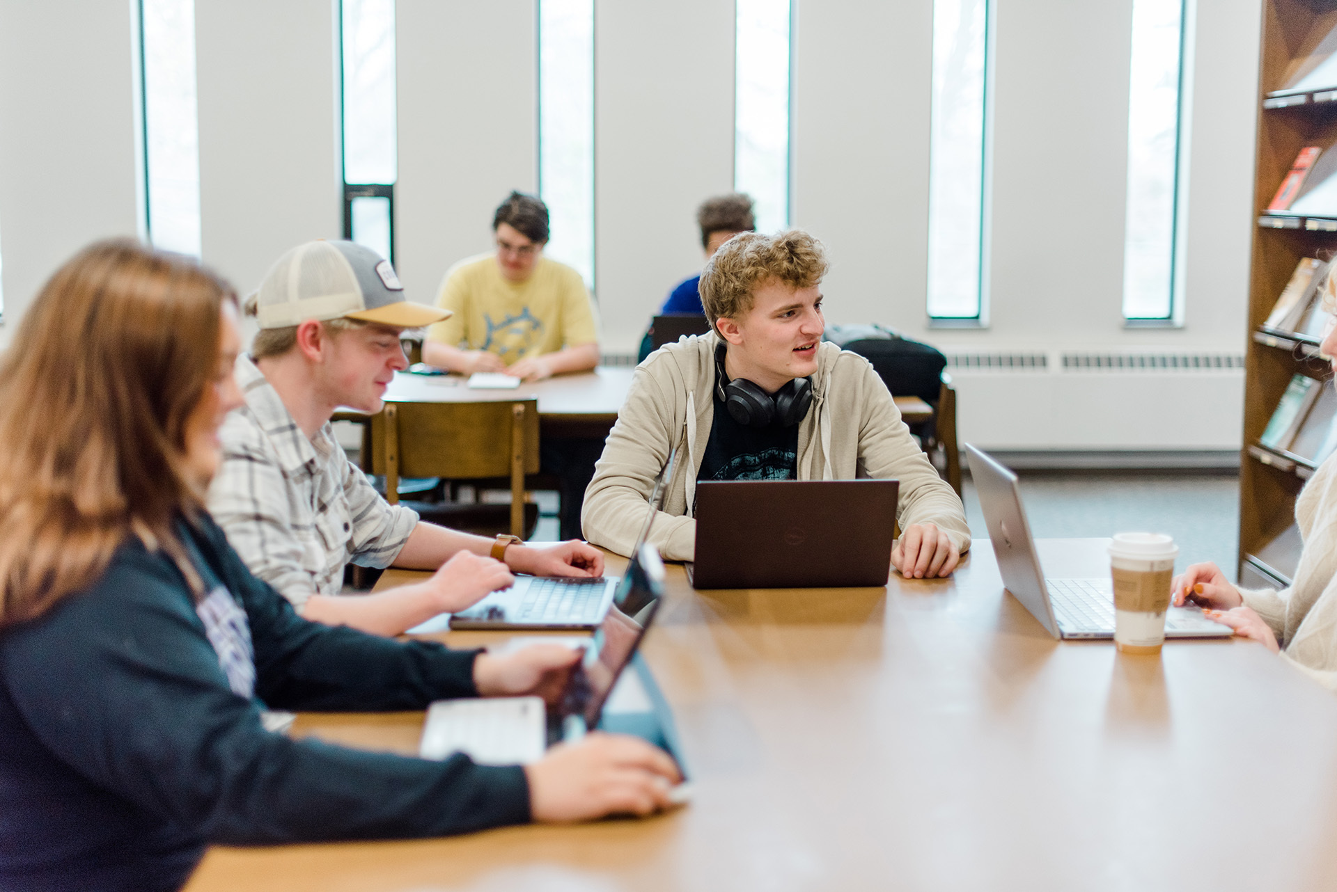 A group of young adults sit around a wooden table in a well-lit library. Some of them are using laptops, and others have notebooks. One person has headphones around their neck and a coffee cup on the table. Bookshelves and large windows are in the background.