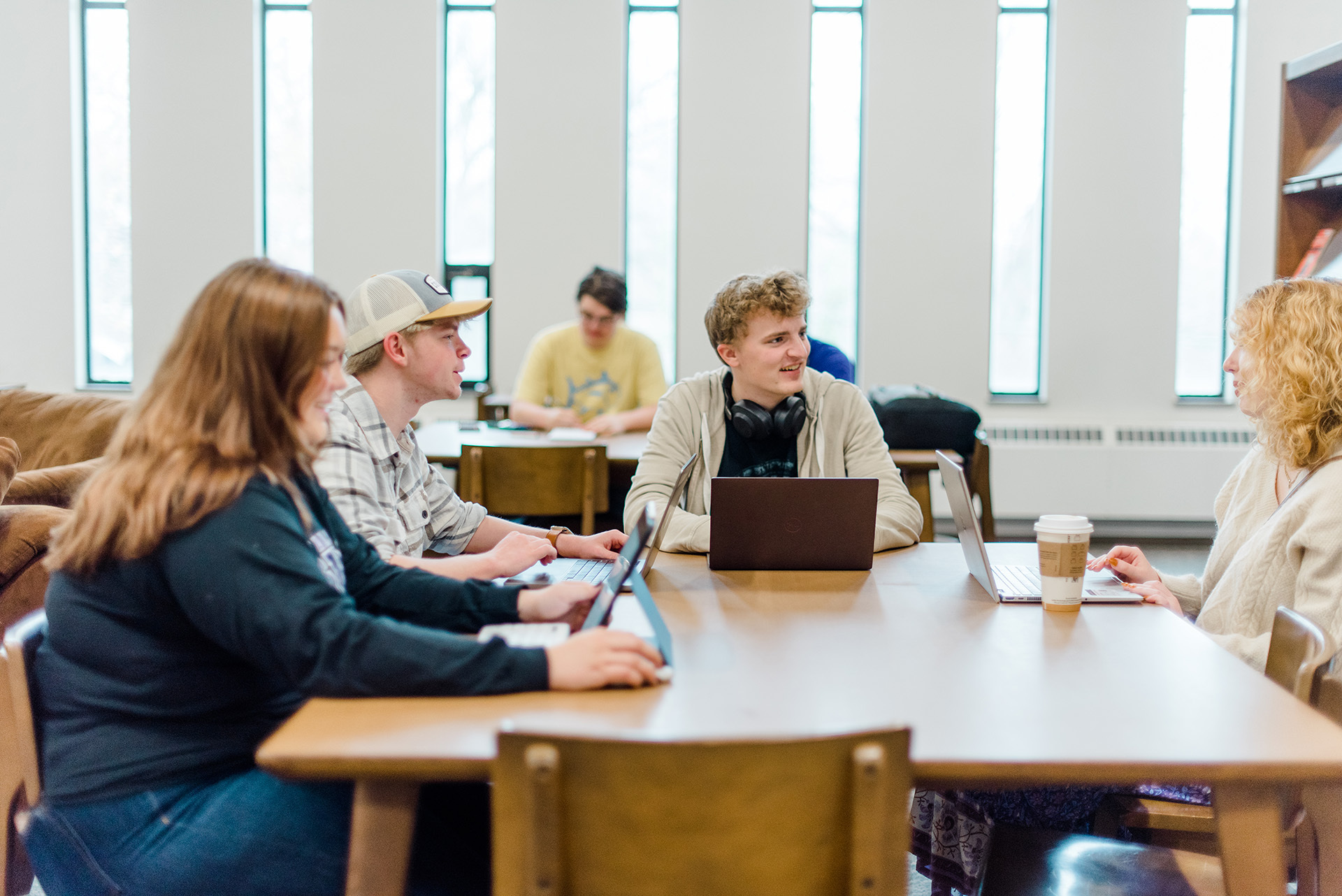 A group of four people are sitting around a wooden table in a brightly lit room with tall windows. They are engaged in a conversation, with laptops and a coffee cup on the table. In the background, another person is working at a separate table.