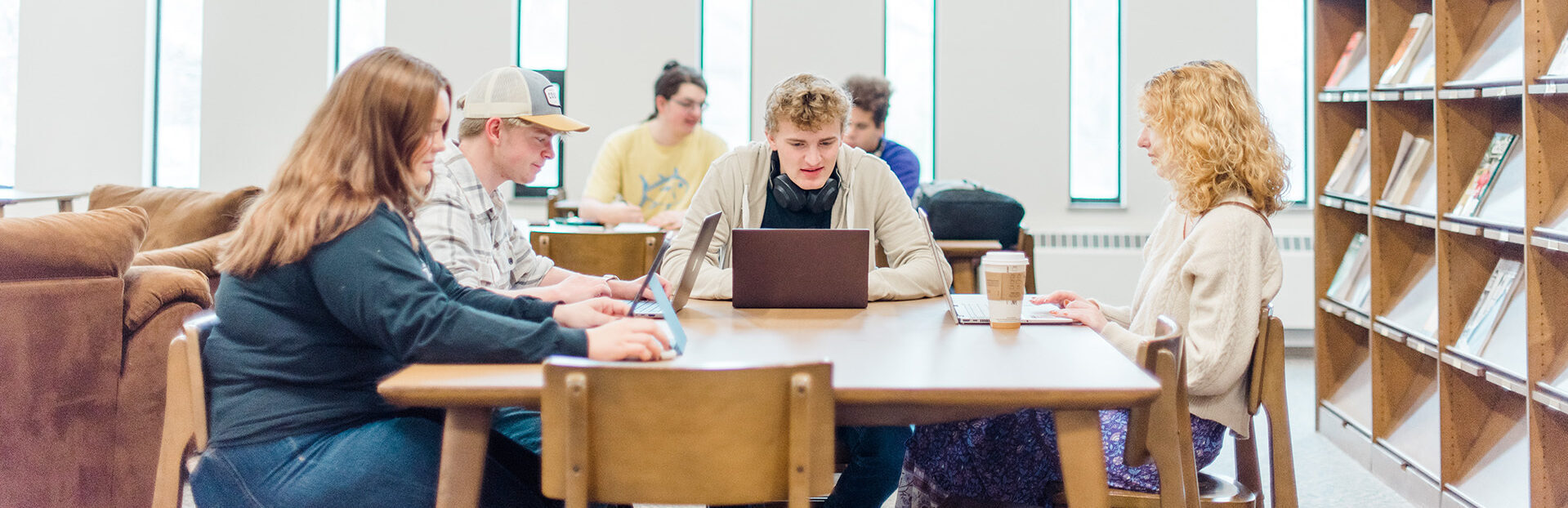 A group of young adults is sitting around a table in a bright library, focused on their laptops and notebooks. Shelves of books and large vertical windows are in the background. The overall atmosphere is one of study and collaboration.