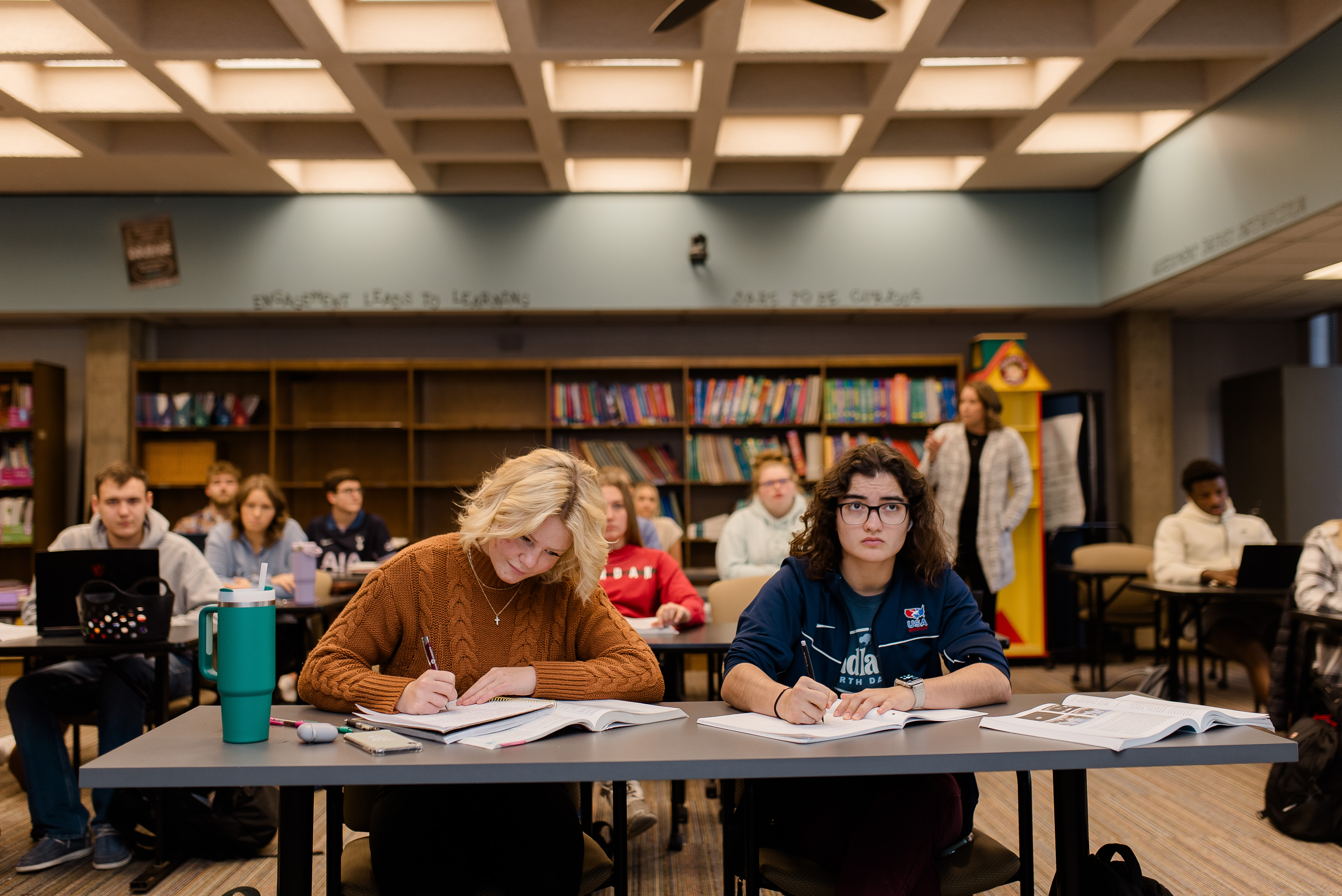 A diverse group of students sits at desks in a classroom, attentively taking notes. The room has bookshelves filled with colorful books and a banner that reads, "Dedicated to Learning." A teacher stands at the back, overseeing the class.