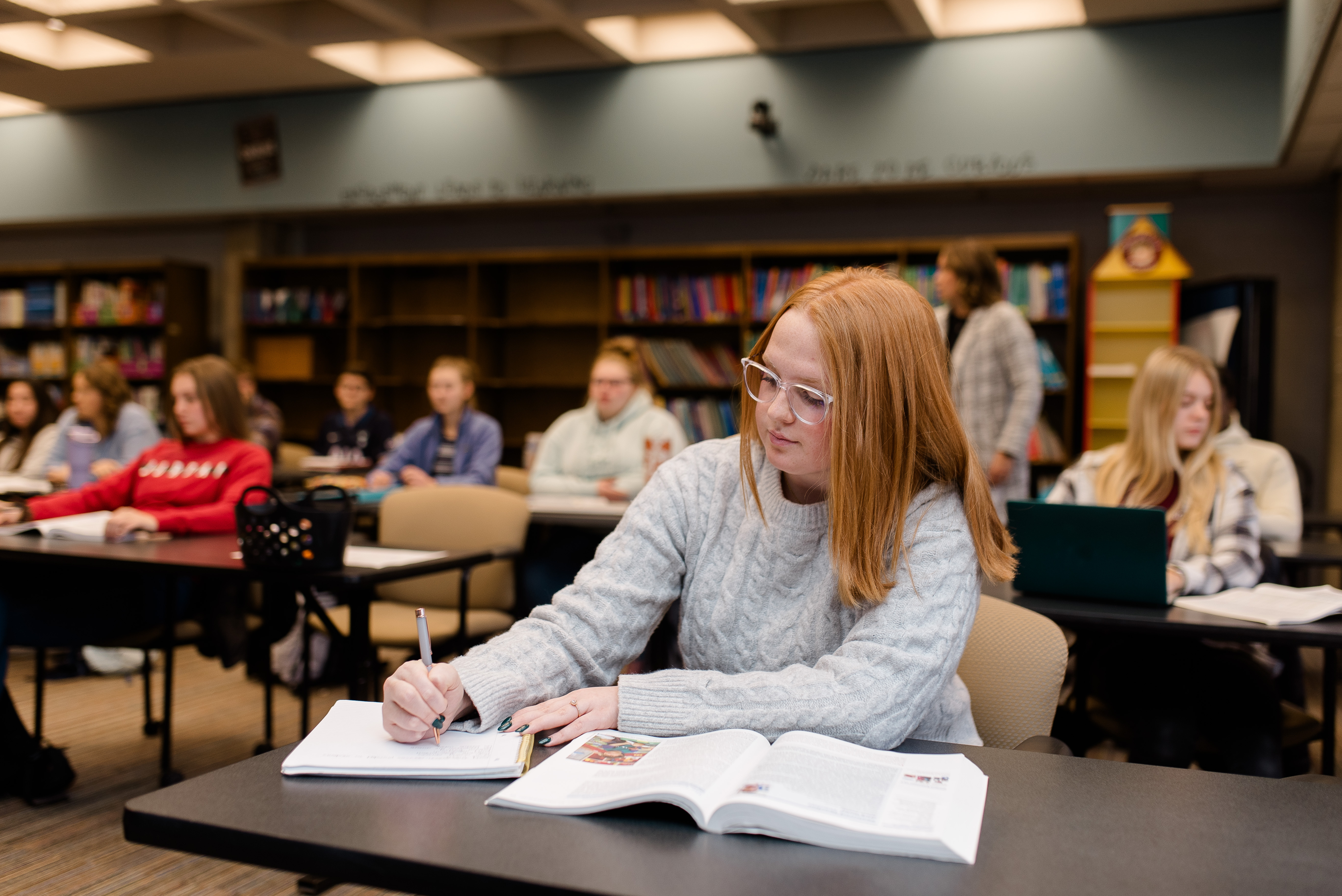 A classroom scene featuring students sitting at tables. In the foreground, a student with red hair and glasses is focused on writing in a notebook, with an open textbook in front of her. Other students in the background are engaged in various activities.