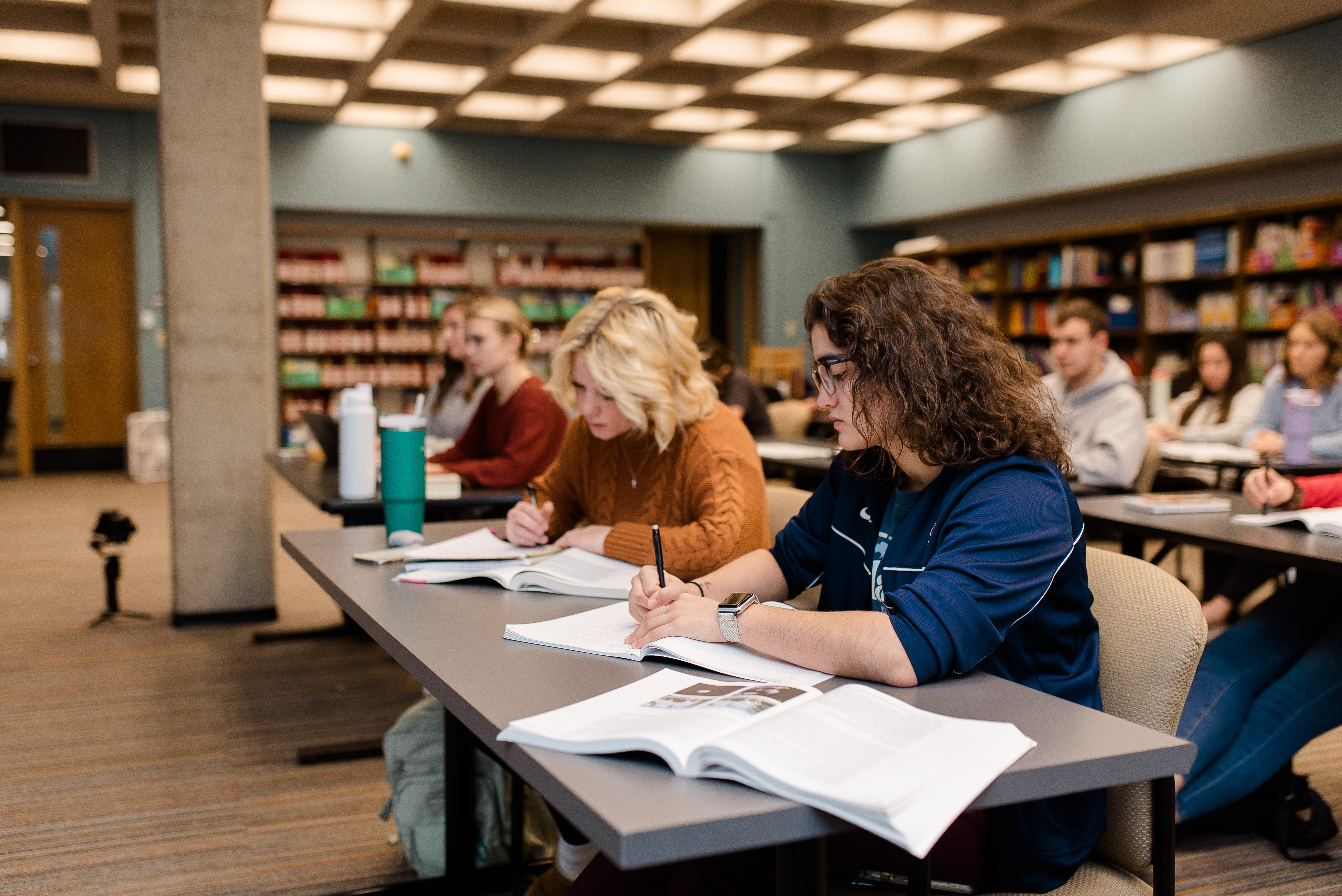 A group of students is seated in a classroom at tables with notebooks and open textbooks. Two students in the foreground are writing intently, while others in the background are focused on their study materials. Bookshelves can be seen lining the back wall.