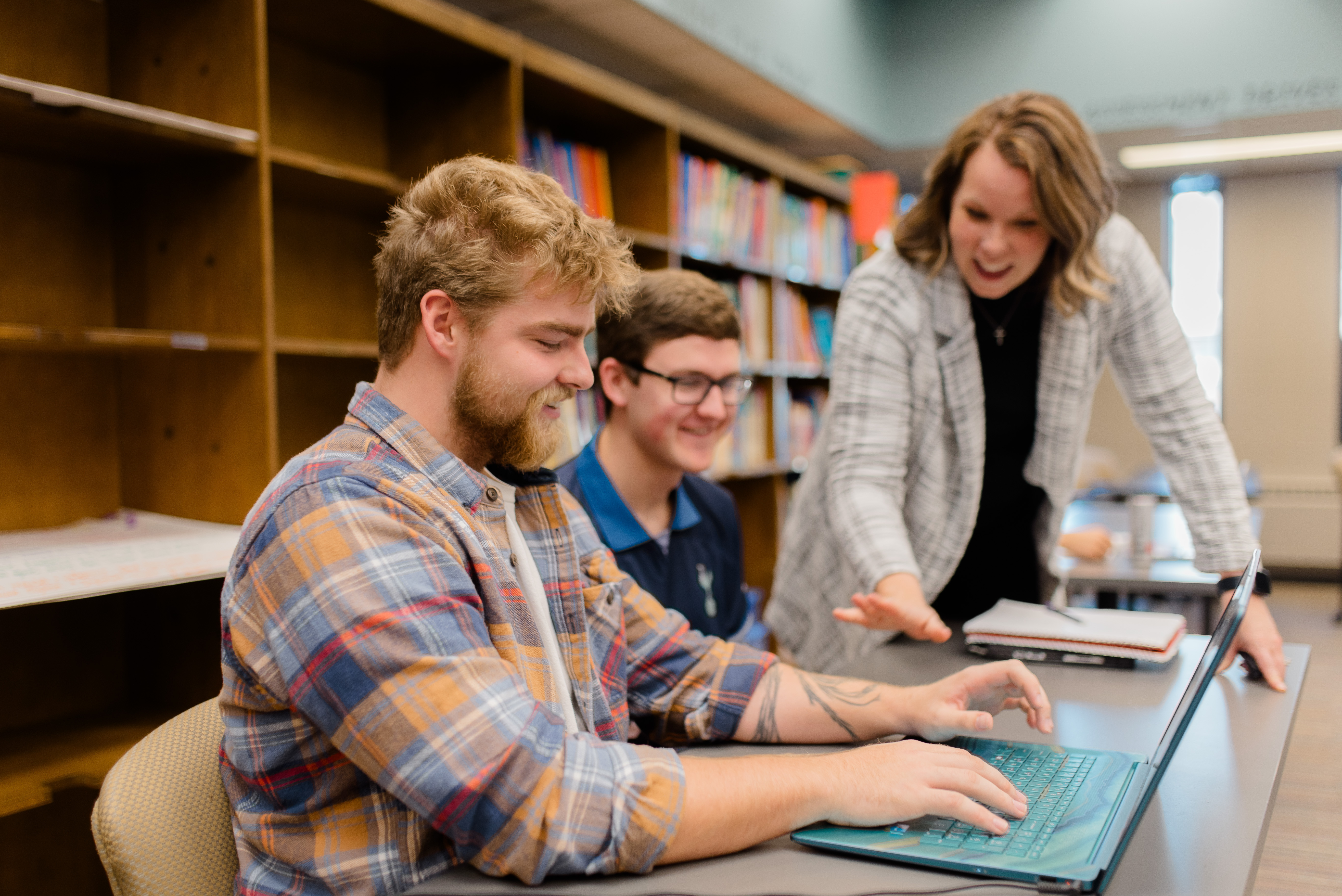 Three people are engaging in a study session. Two young men are seated at a table, focusing on a laptop, with one typing. A woman stands beside them, pointing at the screen and smiling. Bookshelves in the background create a library-like setting.