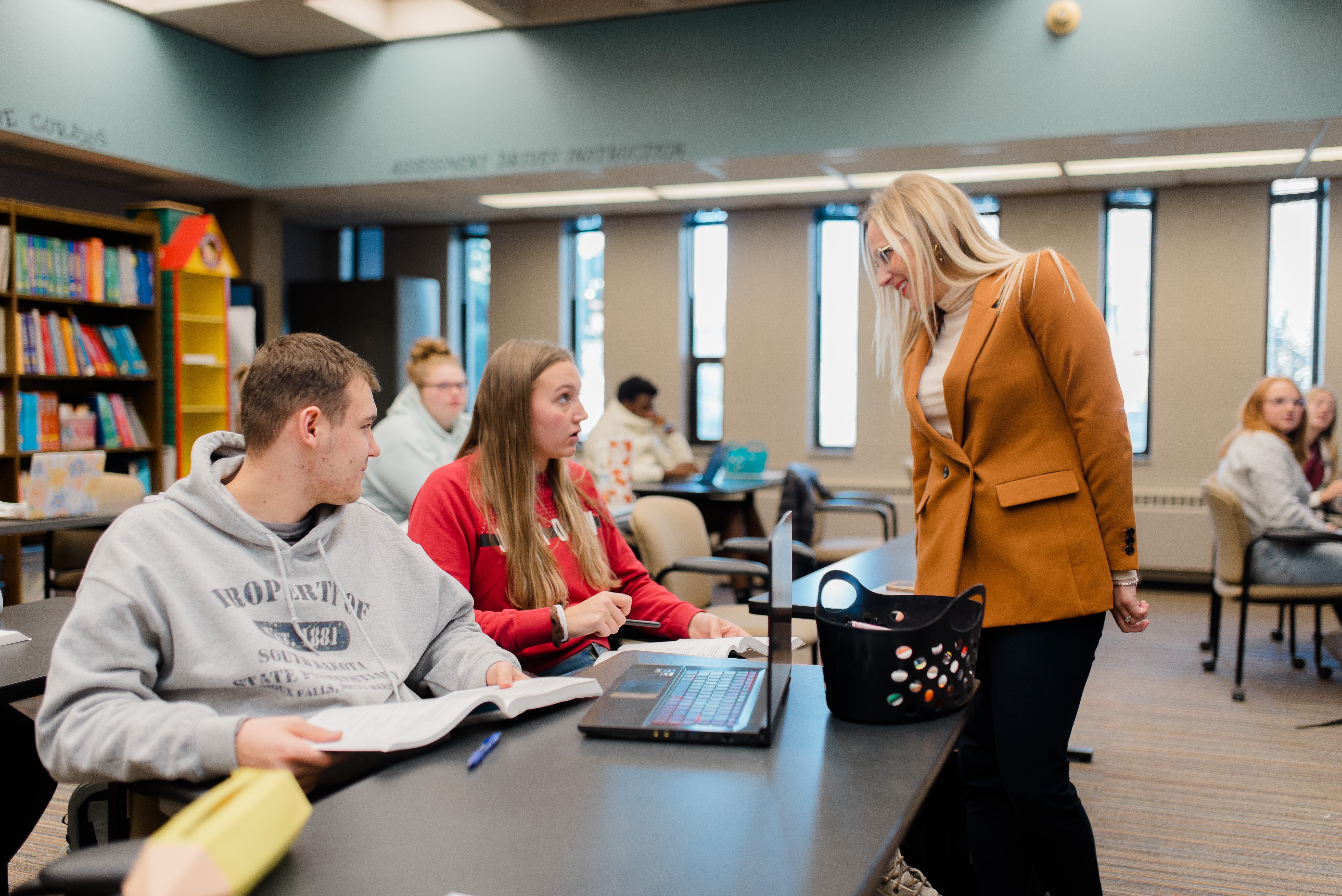 A classroom scene with students sitting at desks, engaging with open books and laptops. A teacher in an orange blazer is leaning over to speak with a female student in a red sweater. Bookshelves and other students are visible in the background.