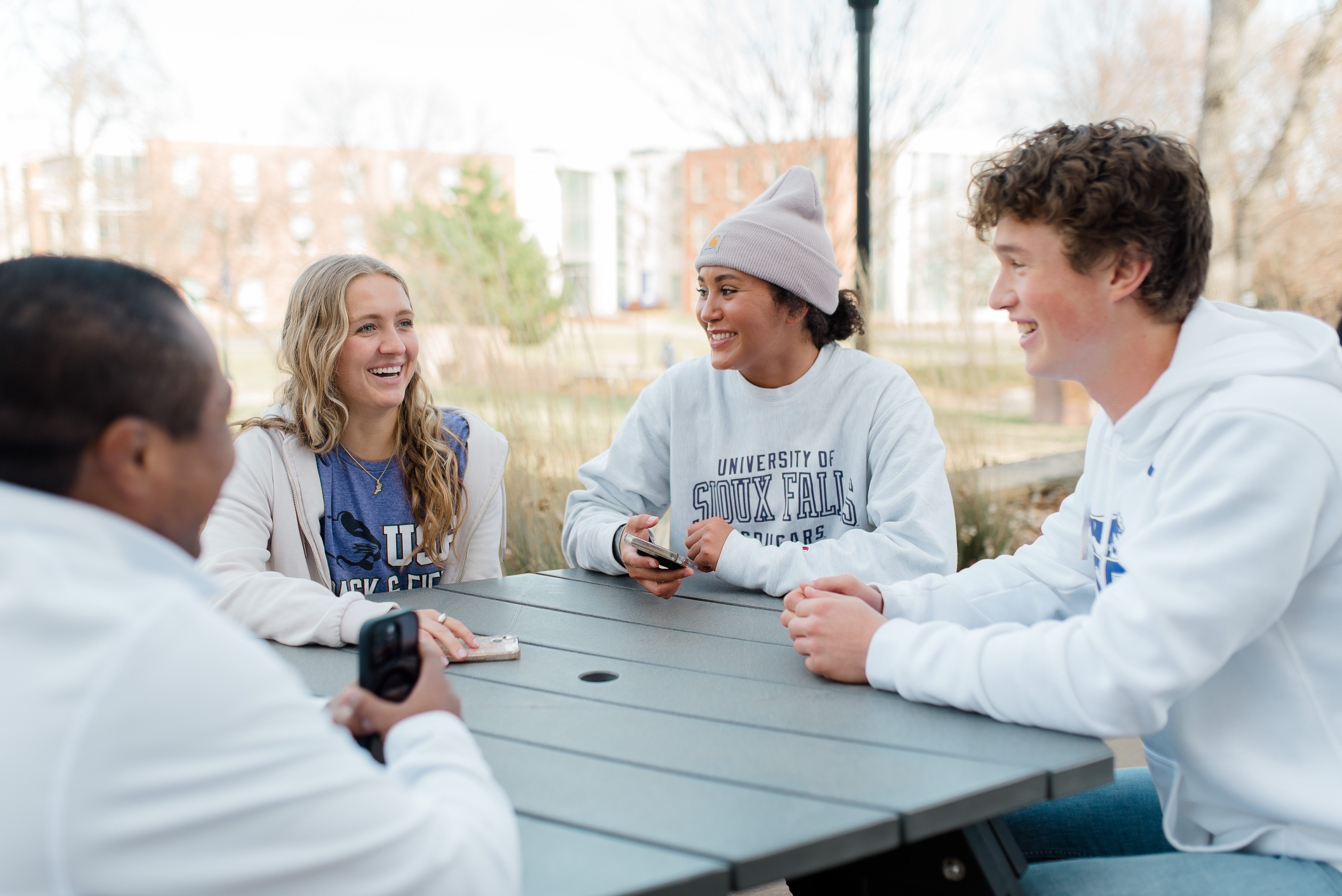 Four young adults sit around an outdoor table on a sunny day. They are smiling and engaged in conversation. Two of them wear white hoodies with printed text, and one wears a beanie. Background includes trees and buildings. They seem to be enjoying each other's company.