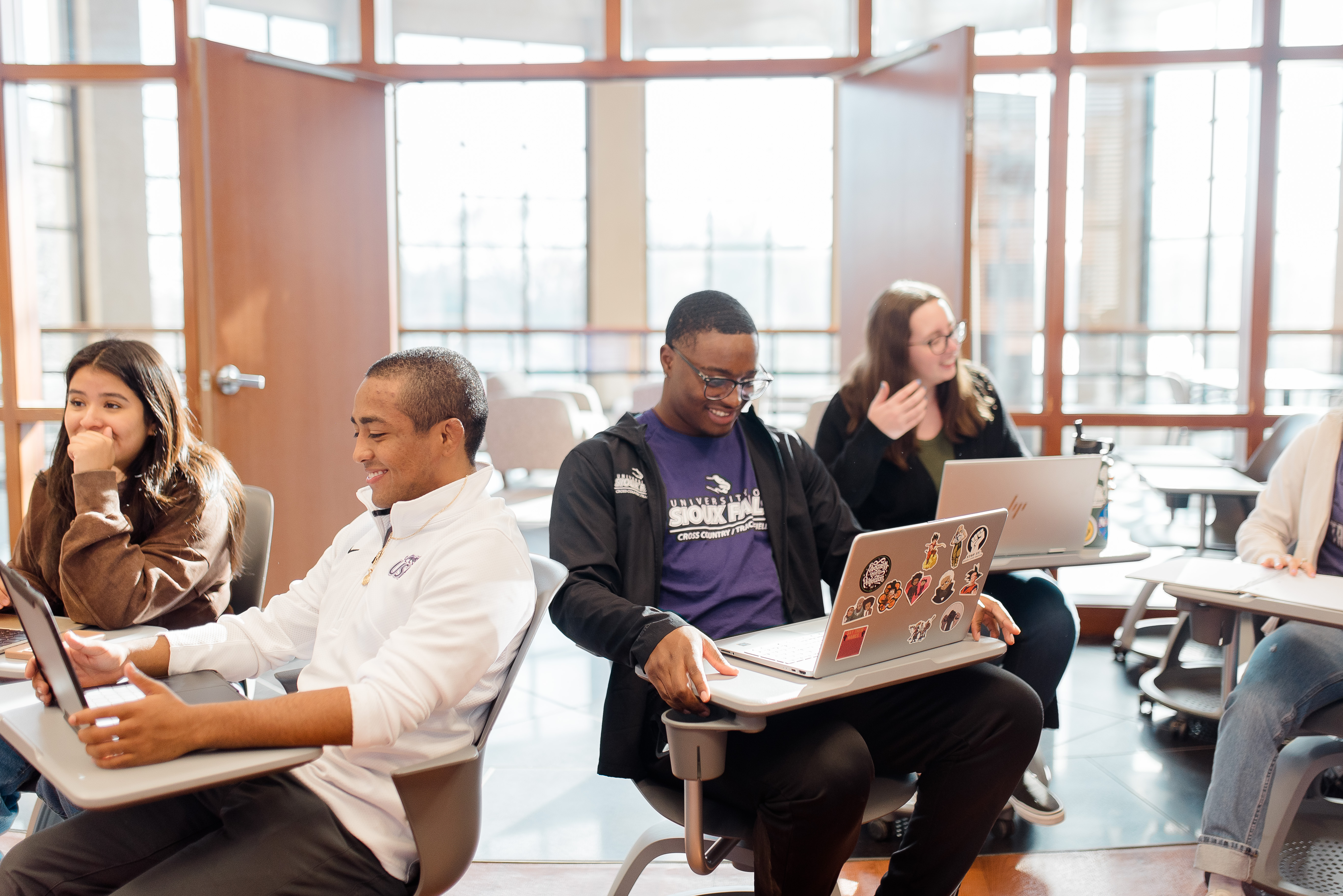 Four students sit at desks with laptops in a bright classroom with large windows. Two students focus on their screens, one in a white jacket and one in black. A third student in purple laughs, while a fourth in a brown jacket talks with them.