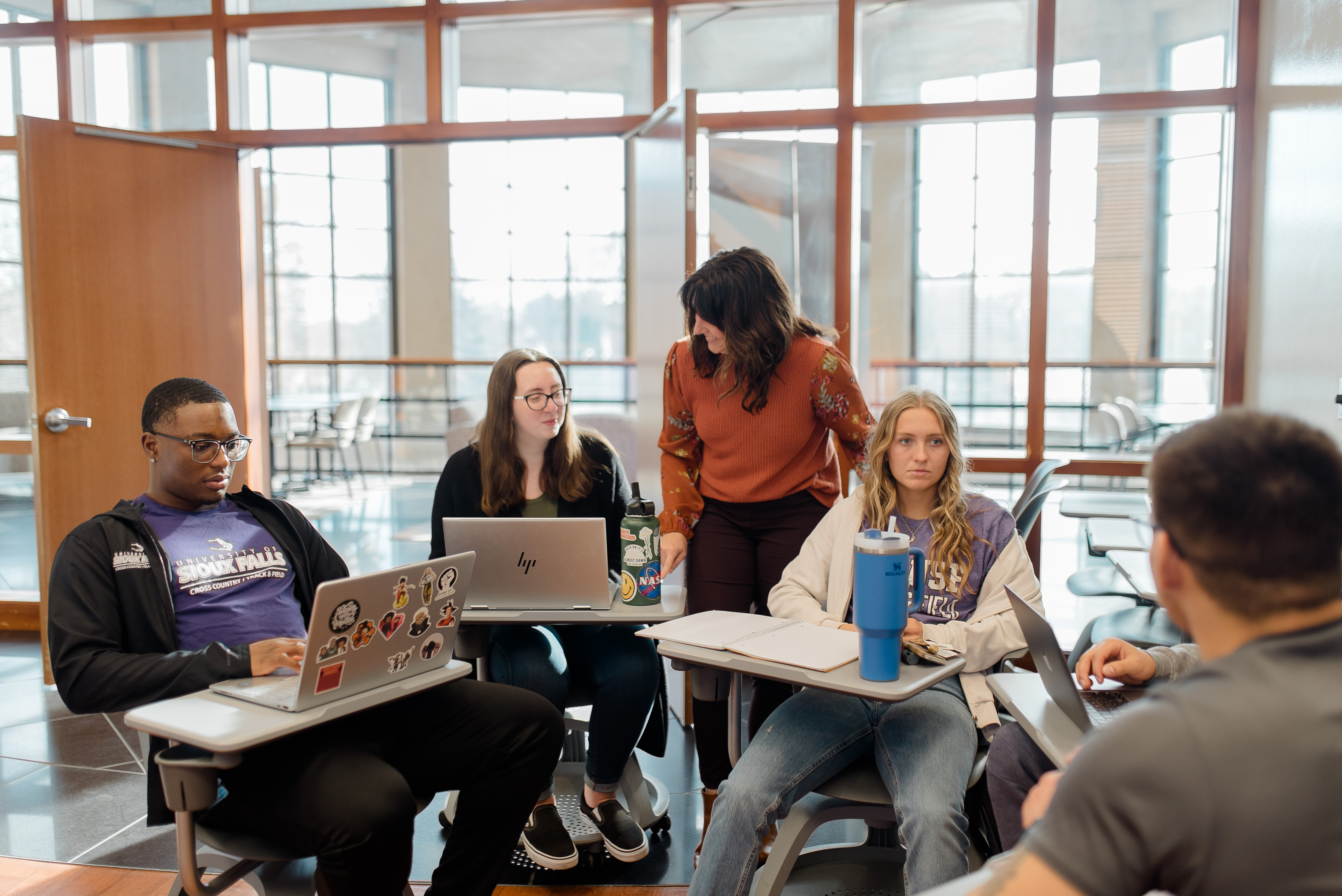 A group of students in a study session inside a modern, well-lit room with large windows. Four students are seated with laptops and notebooks, while one woman stands and engages with them. The atmosphere appears focused and collaborative.