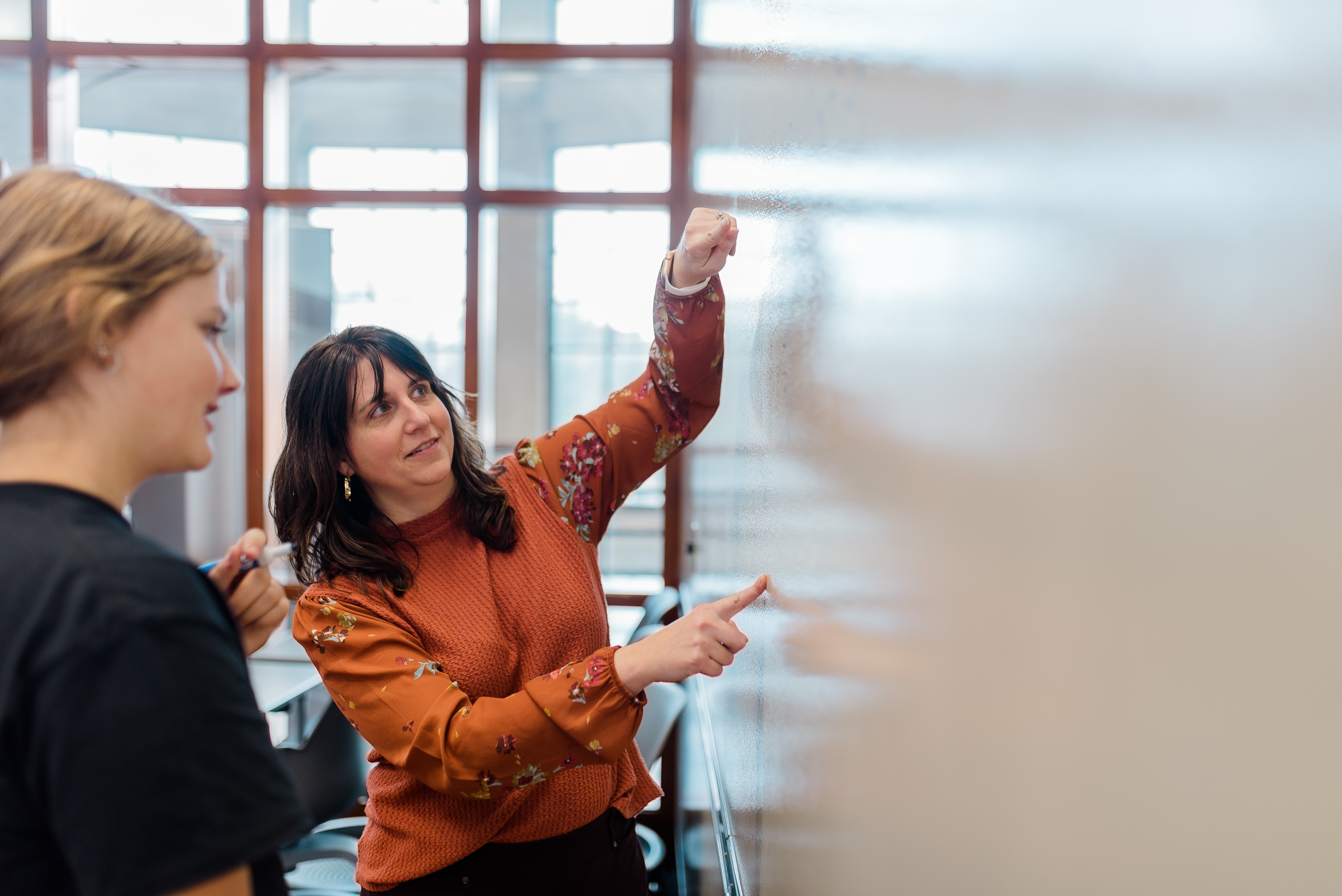 A woman with dark hair in an orange floral blouse points to a whiteboard while talking to a younger woman with light hair in a black shirt. They appear to be in a classroom or meeting room with red-framed windows in the background.