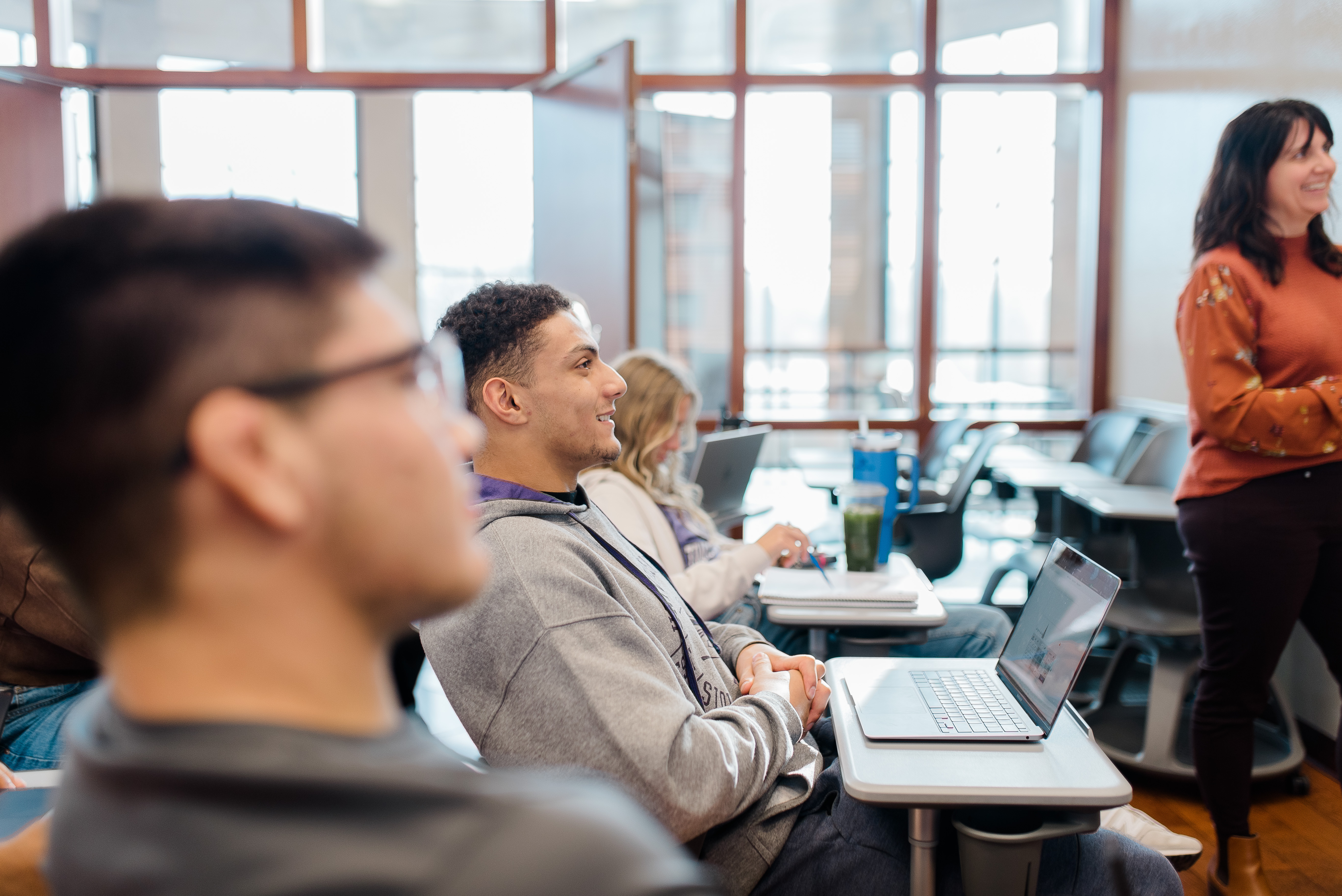 A classroom with students sitting at desks, some using laptops. A young man in the foreground, wearing a hoodie, smiles and looks towards the front. A woman, possibly the teacher, stands at the front, also smiling. Sunlight streams through large windows in the background.