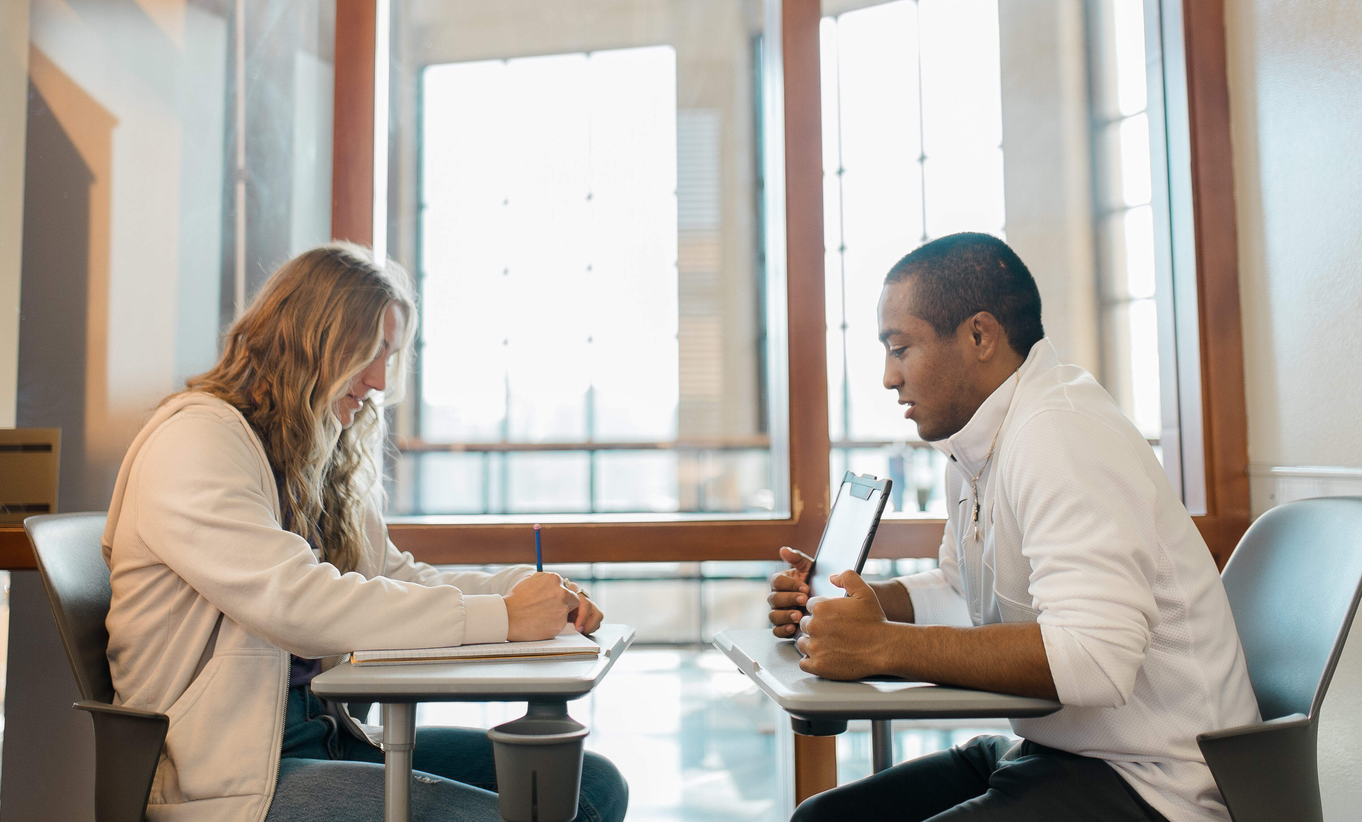 Two people are sitting at a table in a well-lit room. One person is writing in a notebook, while the other is holding a smartphone and engaging in conversation. Both are casually dressed, and the background features large windows with sunlight streaming in.