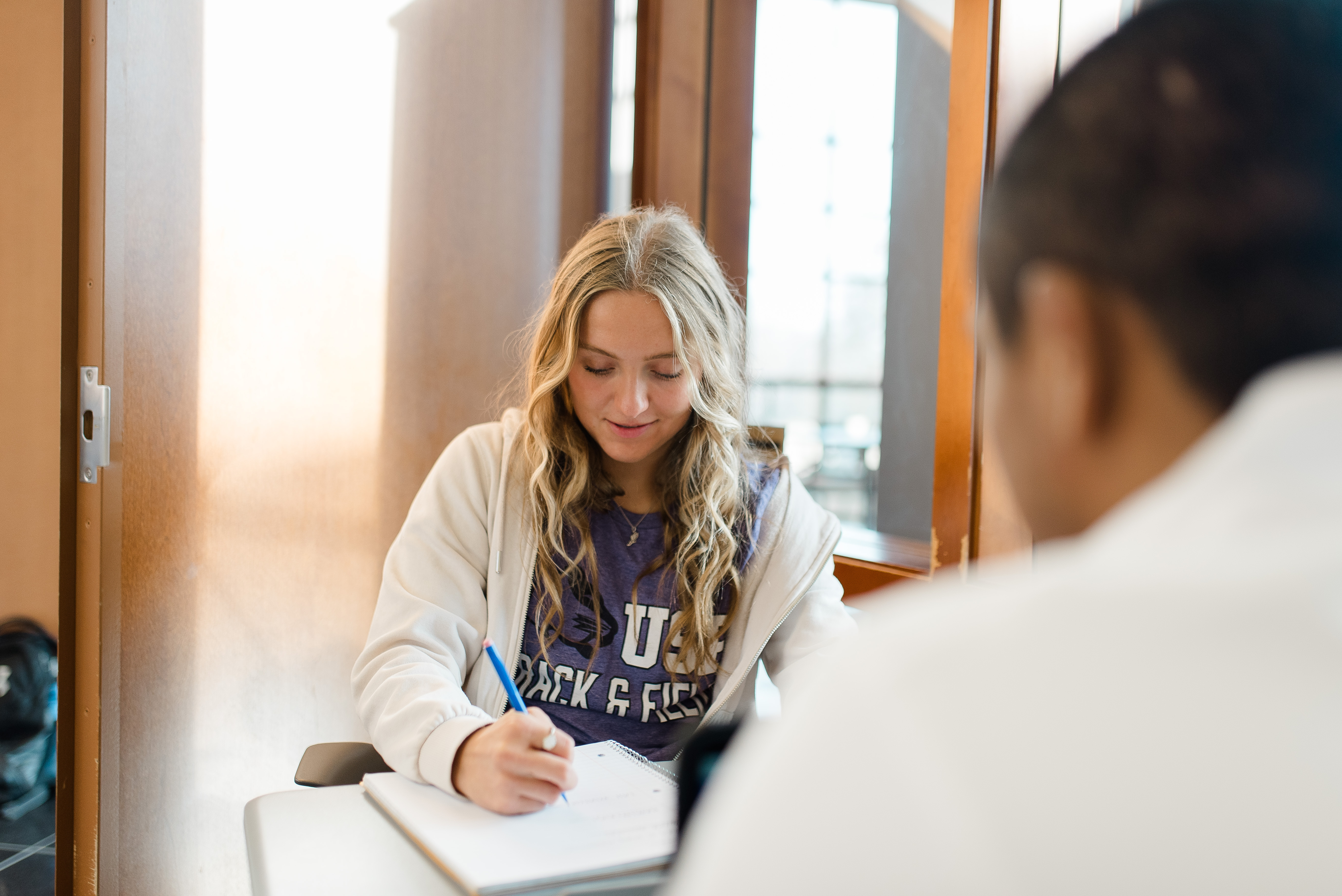 A young woman with long blond hair is seated and writing in a notebook. She is wearing a white jacket over a T-shirt. An out-of-focus person is in the foreground, partially turned toward her. They appear to be interacting in a well-lit indoor space.