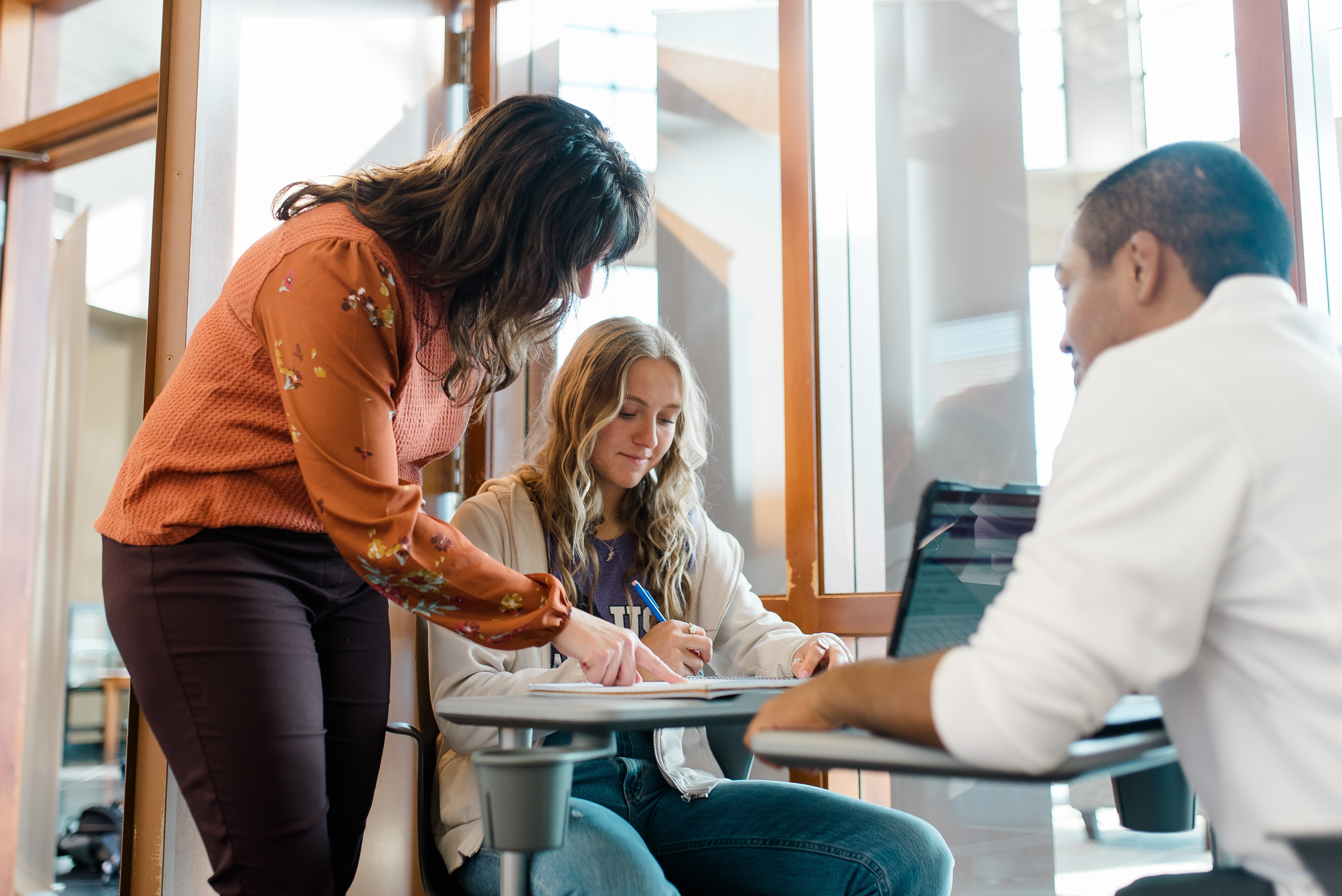 A woman in a rust-orange blouse is leaning over, assisting a seated girl with long blond hair who is writing on a piece of paper. A man in a white shirt sits opposite them, working on a laptop. They are in a bright room with large windows.