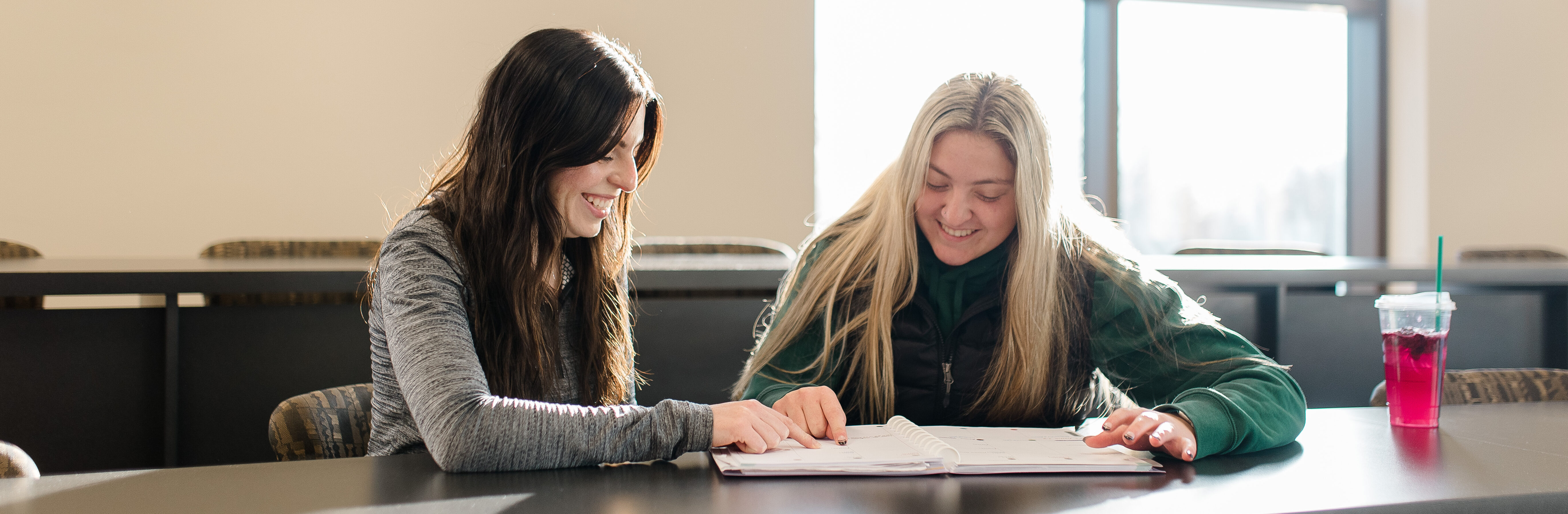 Two young women are sitting at a table in a brightly lit room, smiling and looking at an open notebook together. One has dark hair and the other has blonde hair. A drink with a straw and a pink beverage is placed on the table beside one of them.