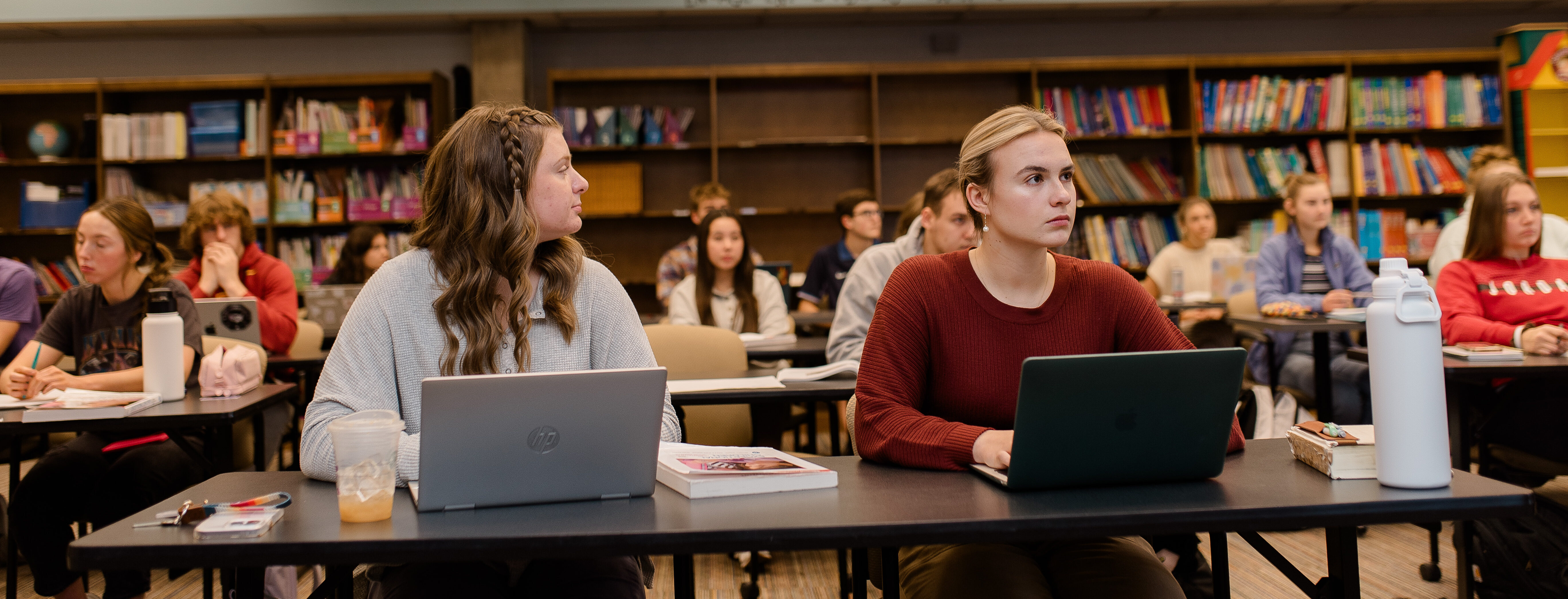 A classroom filled with students sitting at desks, facing forward. Two young women in the foreground are focused on their laptops. Bookshelves filled with colorful books line the back wall. Overhead lights illuminate the room.