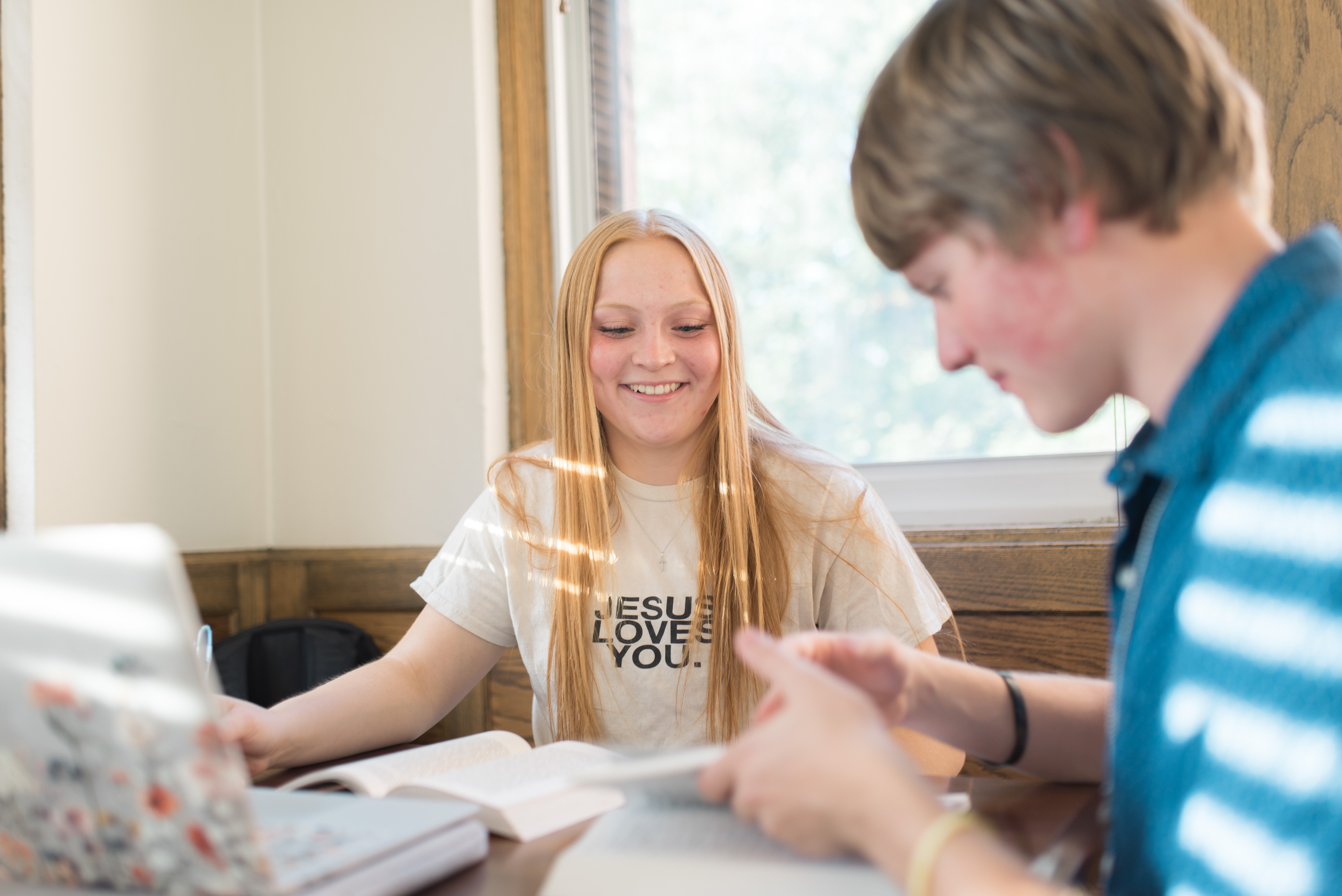 A person with long red hair wearing a white "Jesus Loves You" T-shirt smiles while sitting at a table with books and a laptop. Another person with short blond hair is seated across, focused on a book. Sunlight filters through the window behind them.