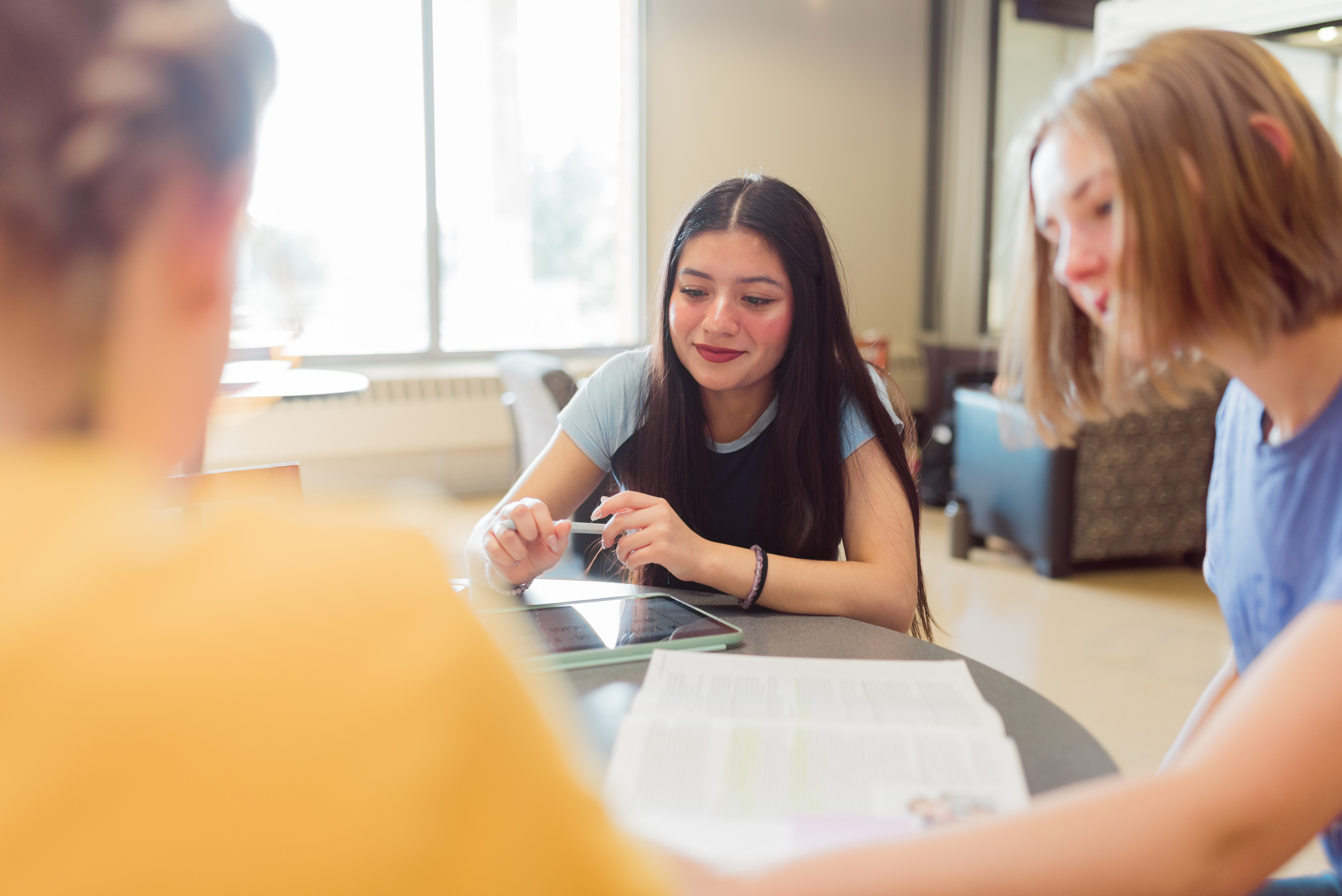 Three young people are sitting at a table indoors with study materials, including books and a tablet. They appear to be engaged in a discussion or group study session. The background is softly lit, with windows and comfortable seating visible.