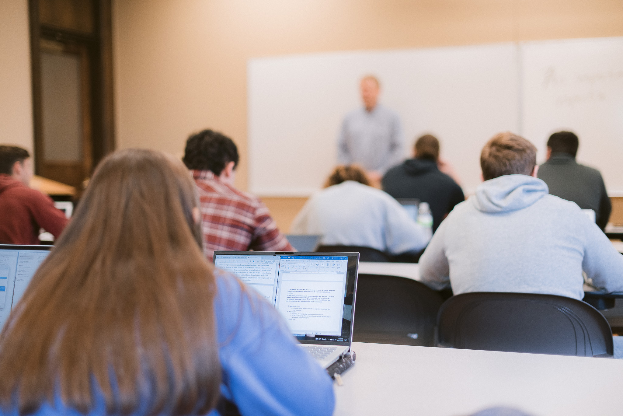 A classroom with students sitting at desks facing a whiteboard. Some students are using laptops; one laptop screen is visible in the foreground. A teacher is standing and talking in front of the class, with a blurred face. The room has a neutral color theme.