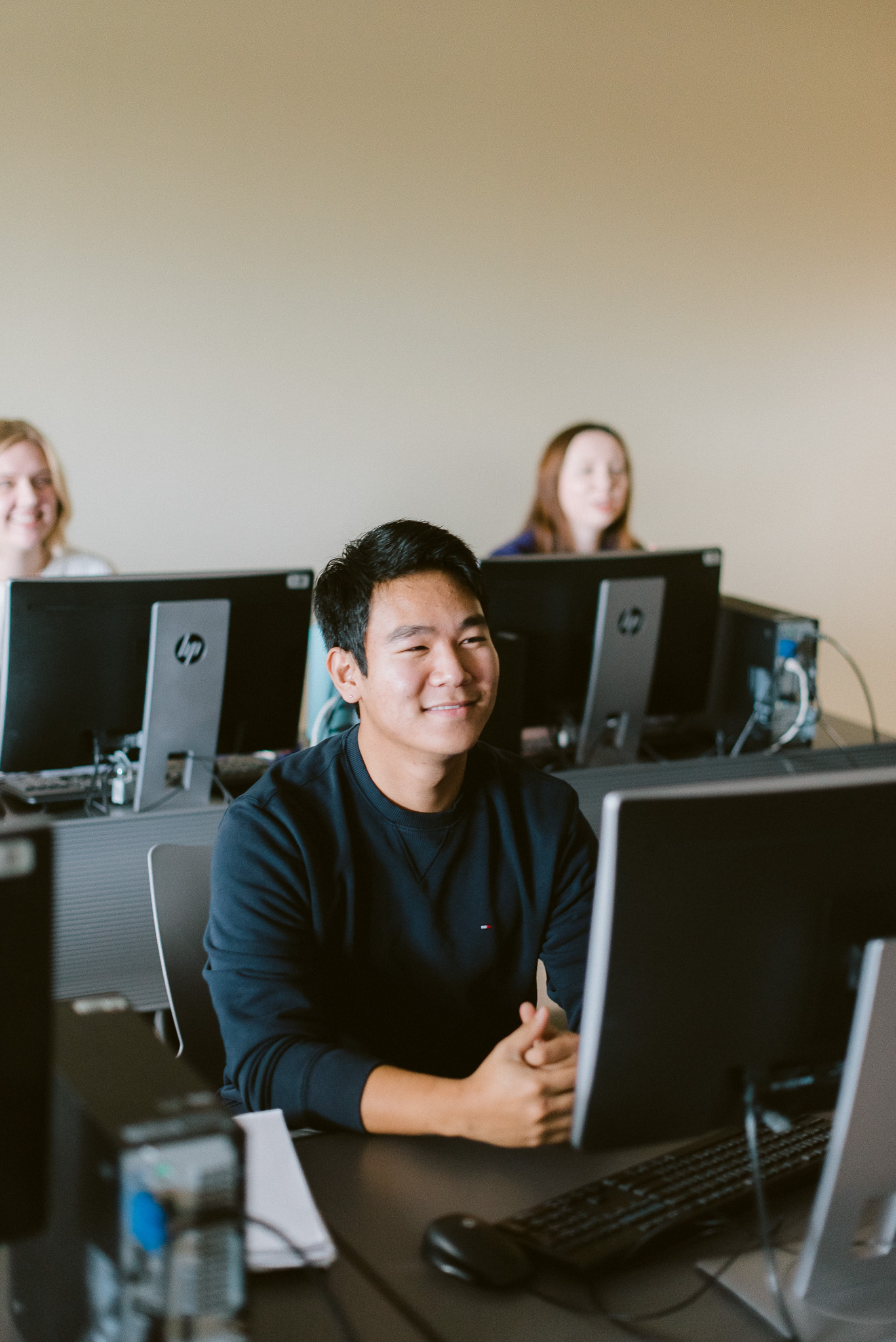 A group of students sit at desks working on desktop computers in a classroom. The central focus is on a smiling individual with short dark hair wearing a black shirt. Two other people are visible in the background, also seated at computers.