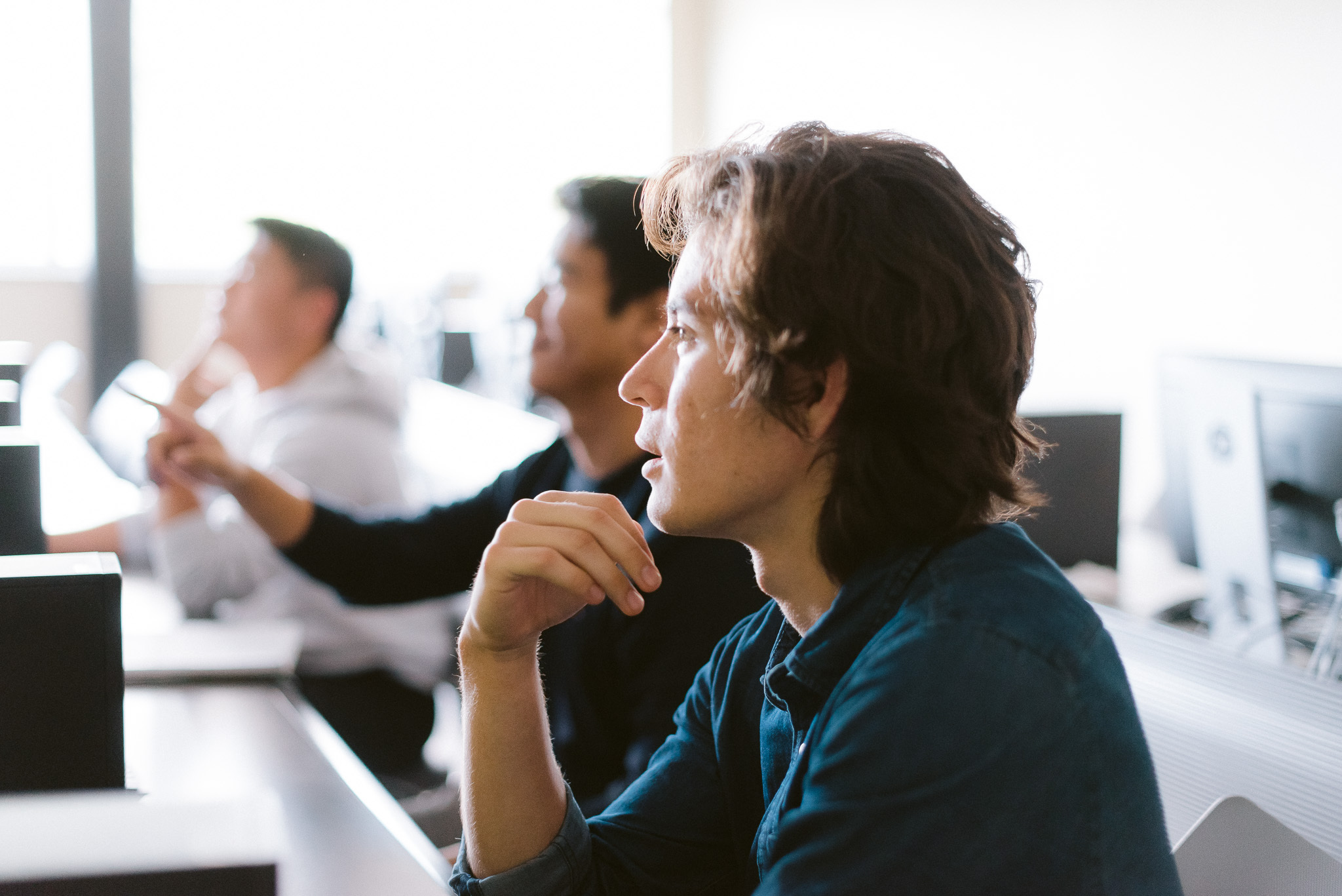 Three people in a classroom setting, each seated at a computer station. The person in focus, in the foreground, is staring intently at a screen with their hand on their chin. The other two people appear slightly blurred in the background.