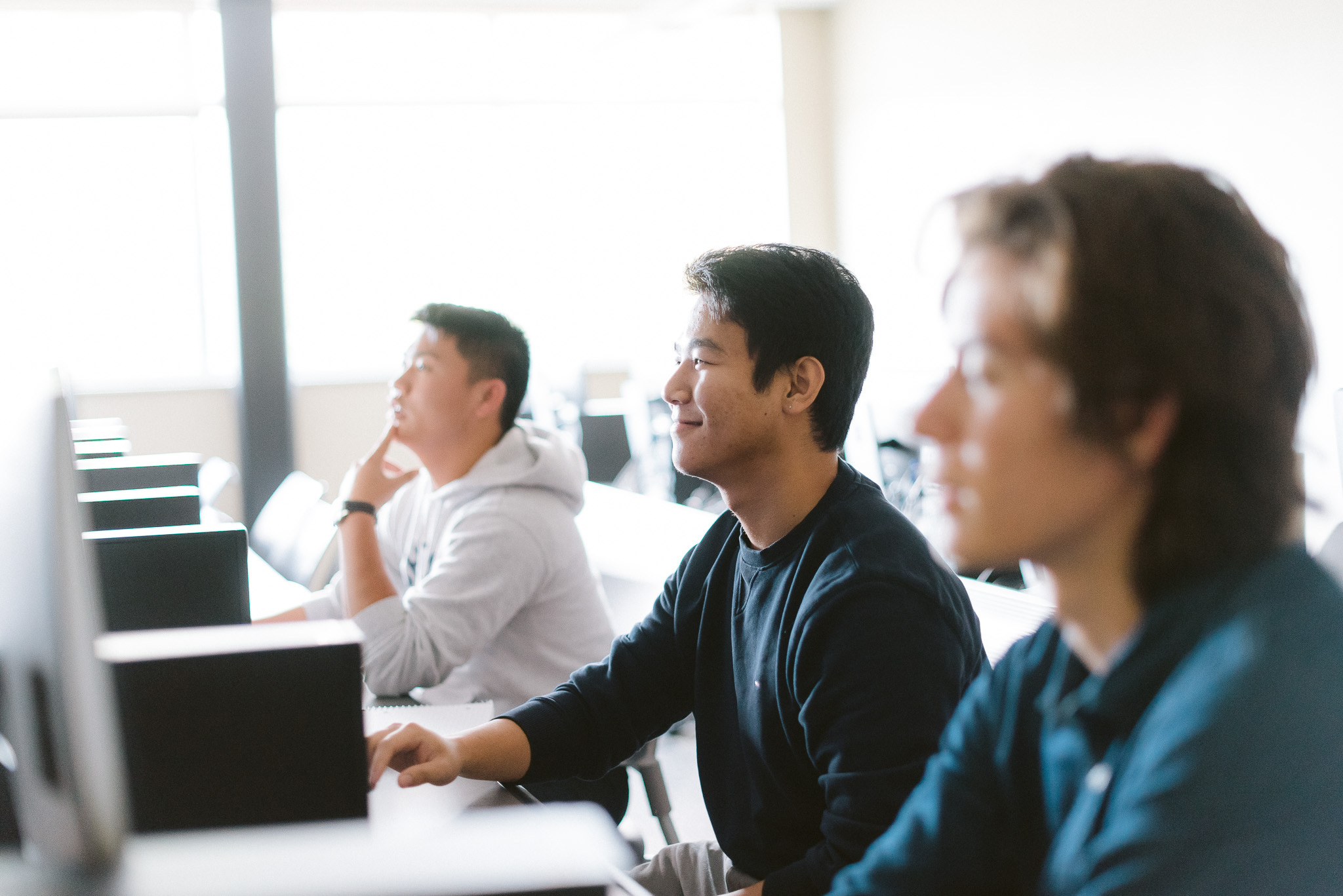 Three students are seated in a row at computer workstations in a classroom. They are focused on their screens and appear engaged in their work. The classroom is bright and has a calm ambiance.