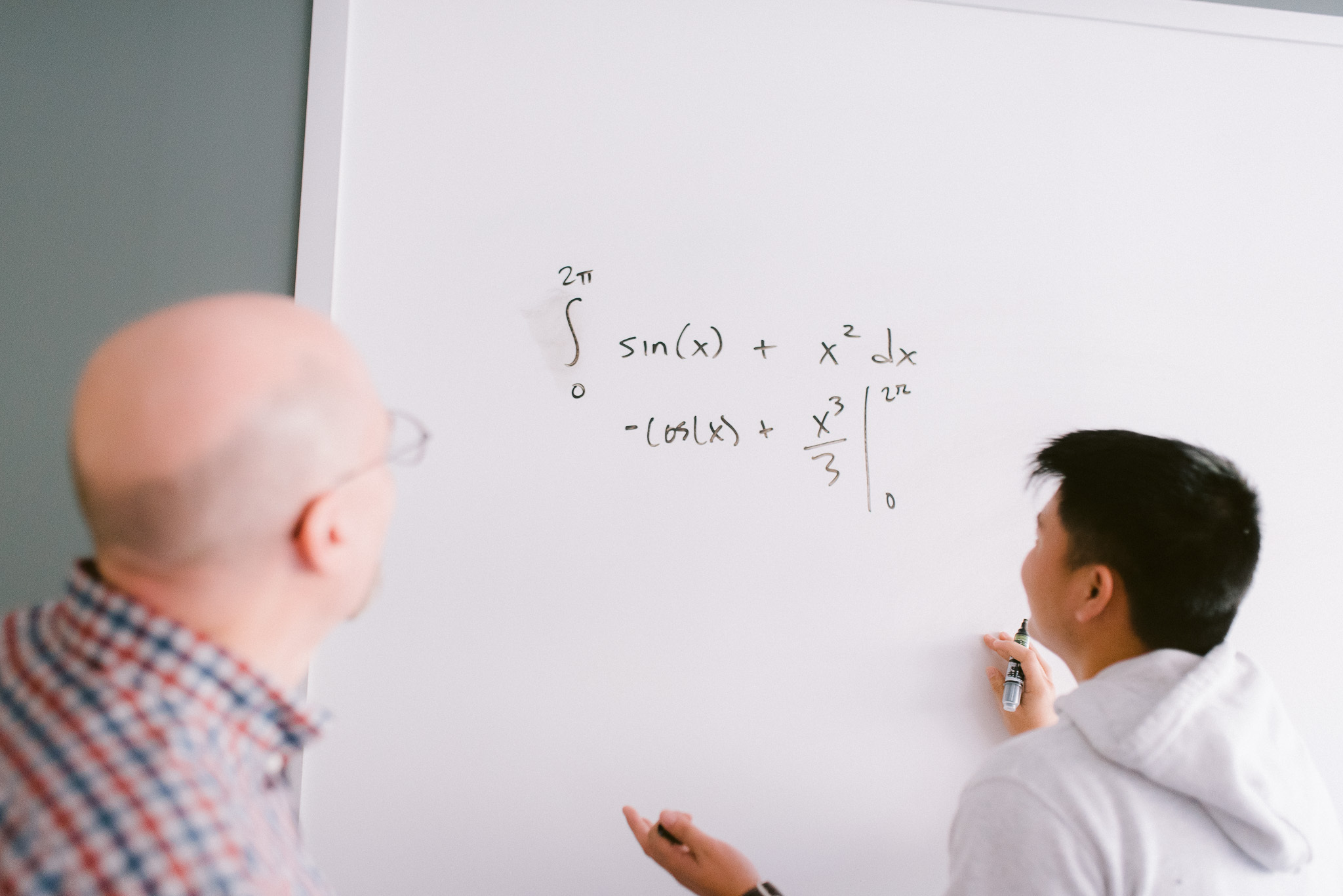 Two people are solving a mathematical integral equation on a whiteboard. One person is drawing on the whiteboard with a marker, while the other observes. The equation includes trigonometric and polynomial functions.