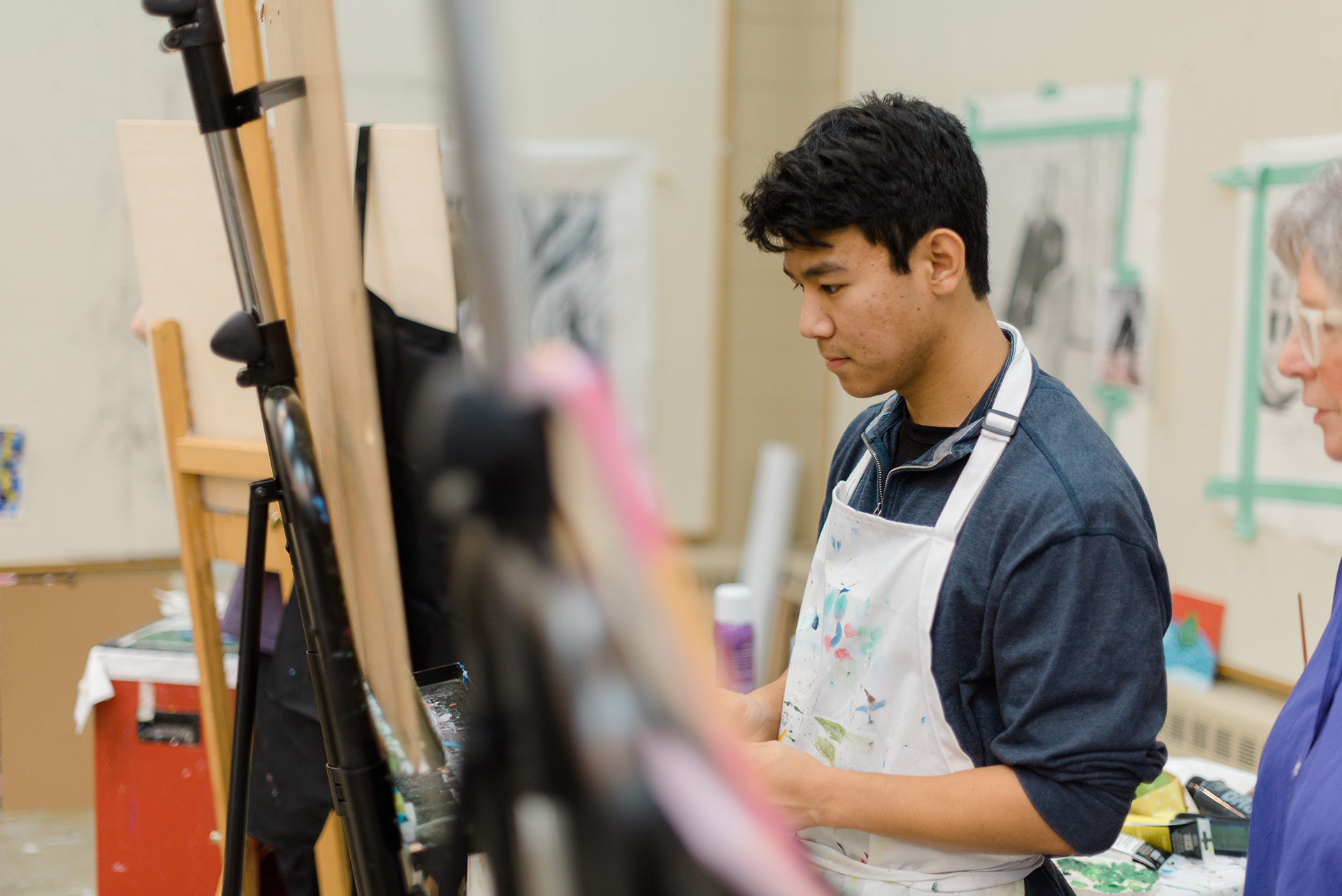 A young man wearing an apron is painting at an easel in an art studio. A woman with gray hair, also in an apron, stands nearby. The studio features paintings and art supplies in the background.