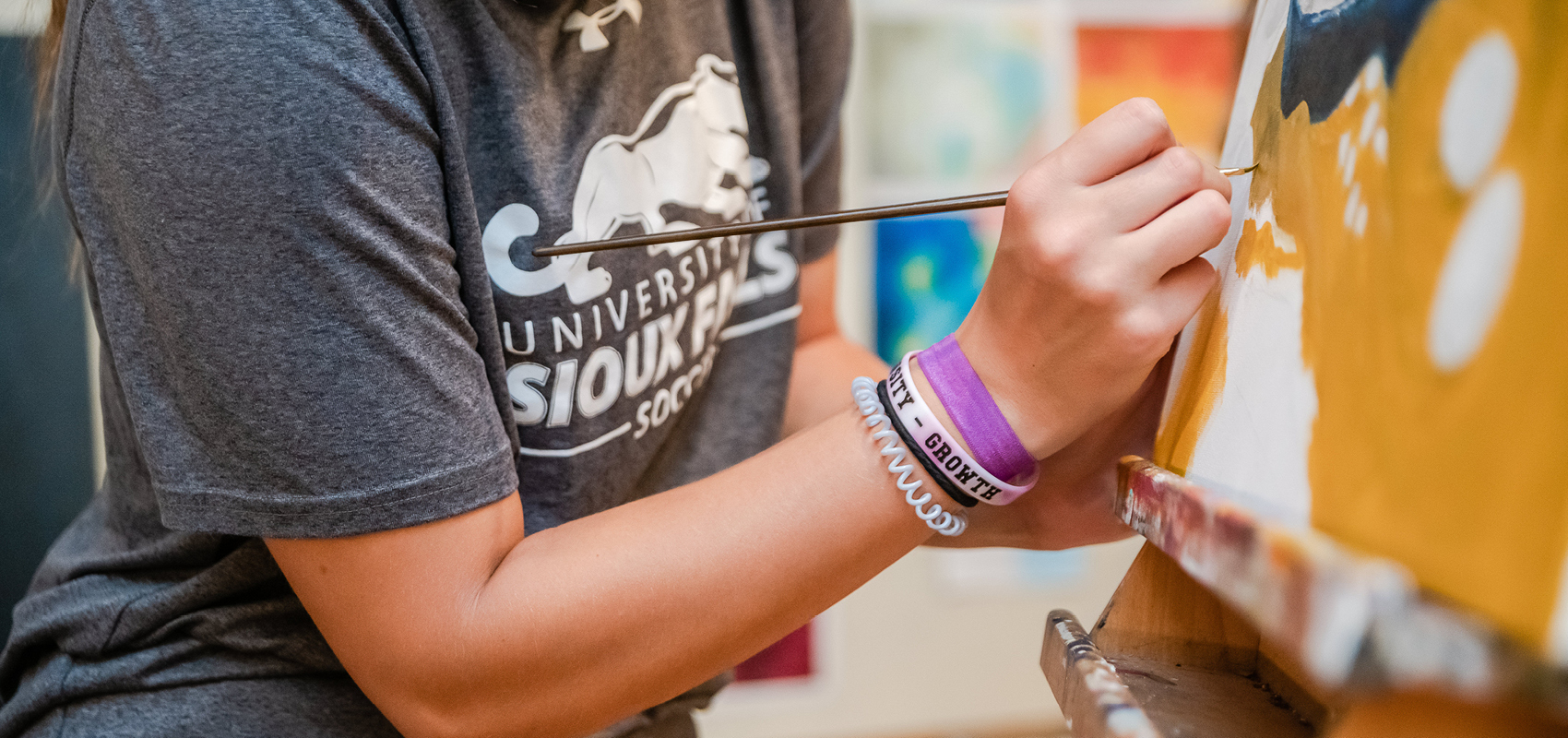 A person in a grey "University of Sioux Falls Soccer" T-shirt is painting on a canvas with a thin brush. They are wearing purple and white bracelets on their wrist. The image is focused on their arm and hand as they create brush strokes on the canvas.
