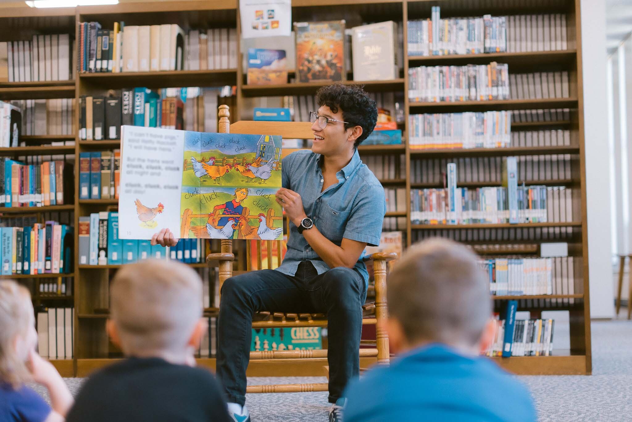 A person sits on a wooden chair in a library, holding up an open picture book and reading to a group of young children sitting on the floor in front. The bookshelves behind them are filled with books and DVDs.