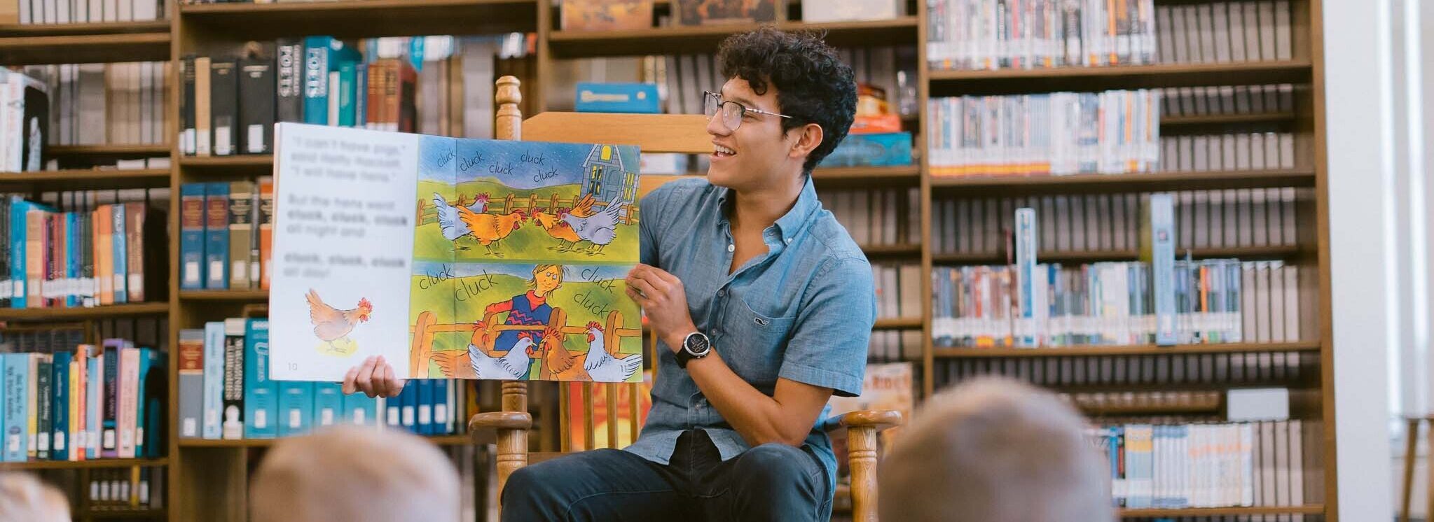 A person with curly hair, wearing glasses and a blue shirt, sits in a wooden chair reading a colorful illustrated book to a group of children in a library. Shelves filled with books are visible in the background.
