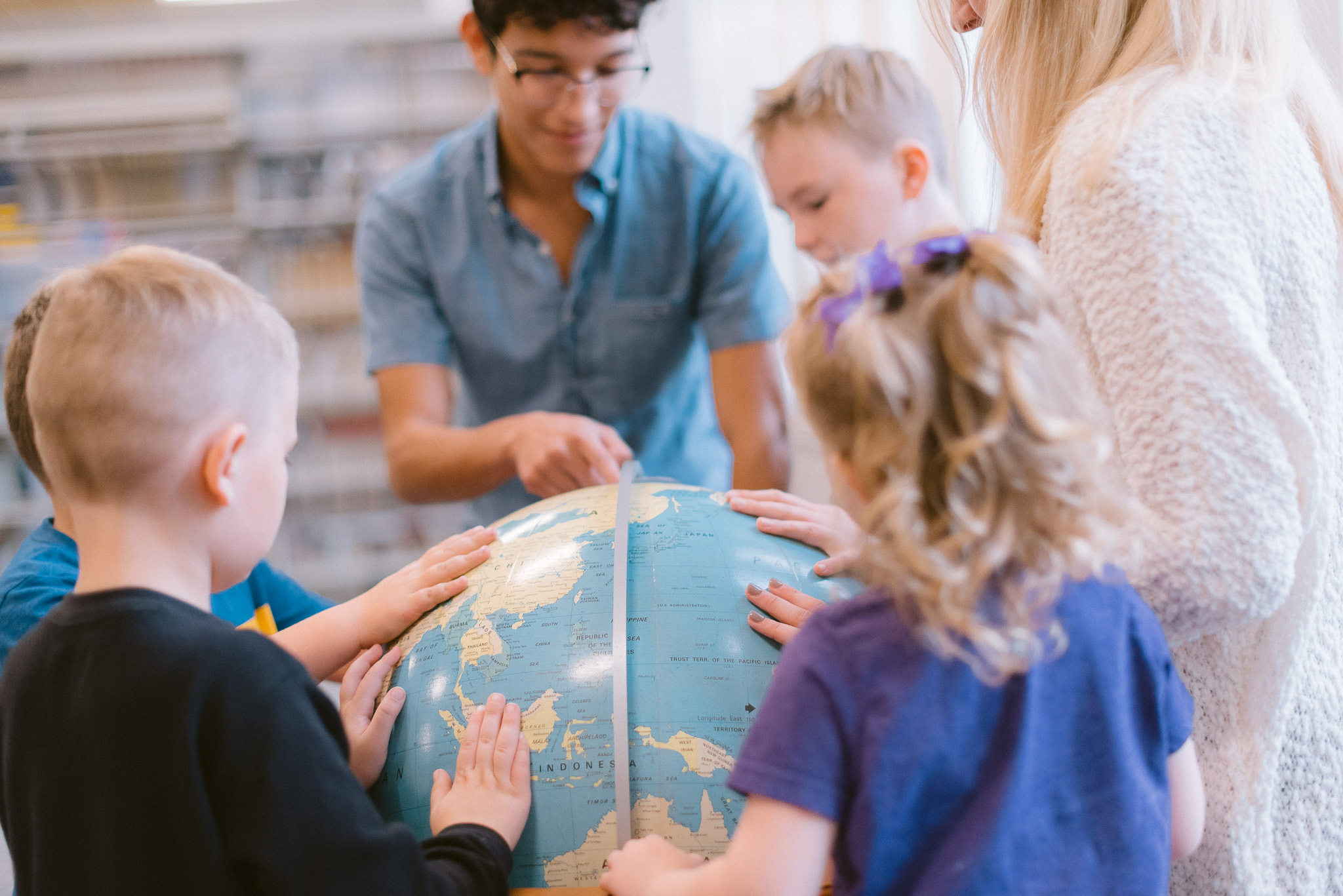 A group of young children gather around a large globe with an adult. They are touching and pointing at various parts of the globe as the adult helps guide their learning.