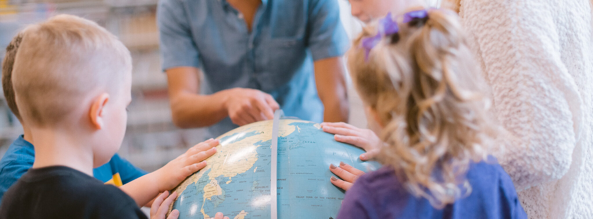 A group of children and an adult gather around a globe. The children place their hands on the globe, and the adult points to a location on it. The scene appears to be educational, possibly in a classroom or learning environment.