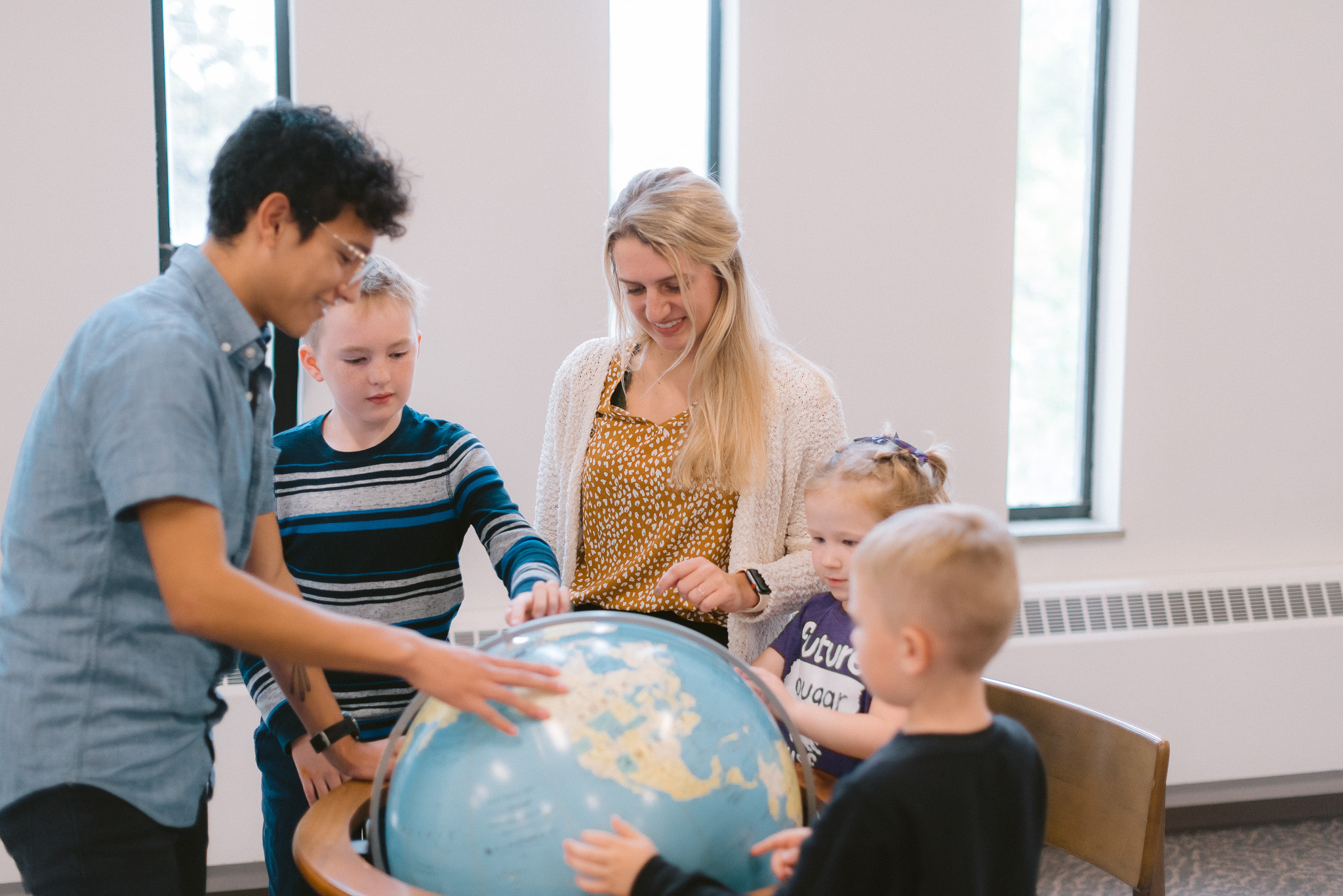 A group of four children and one adult gather around a large globe in a brightly lit room. The children touch the globe while the adult, standing nearby, attentively observes and guides them. Two tall windows with daylight streaming through are in the background.