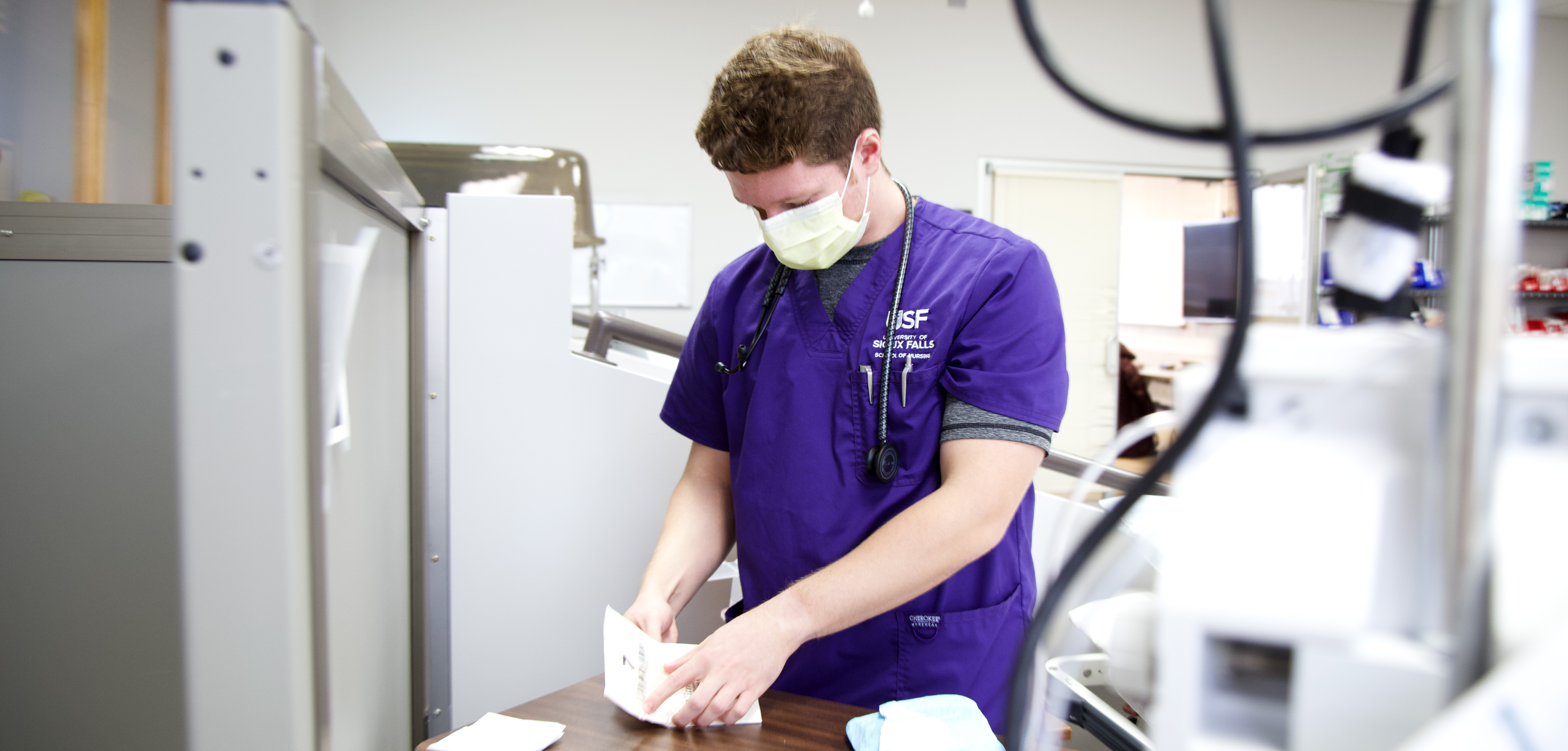 A person wearing a purple medical uniform and a face mask is preparing medical supplies at a workstation. Medical equipment and supplies are visible around the workspace. The individual is focused on the task at hand.