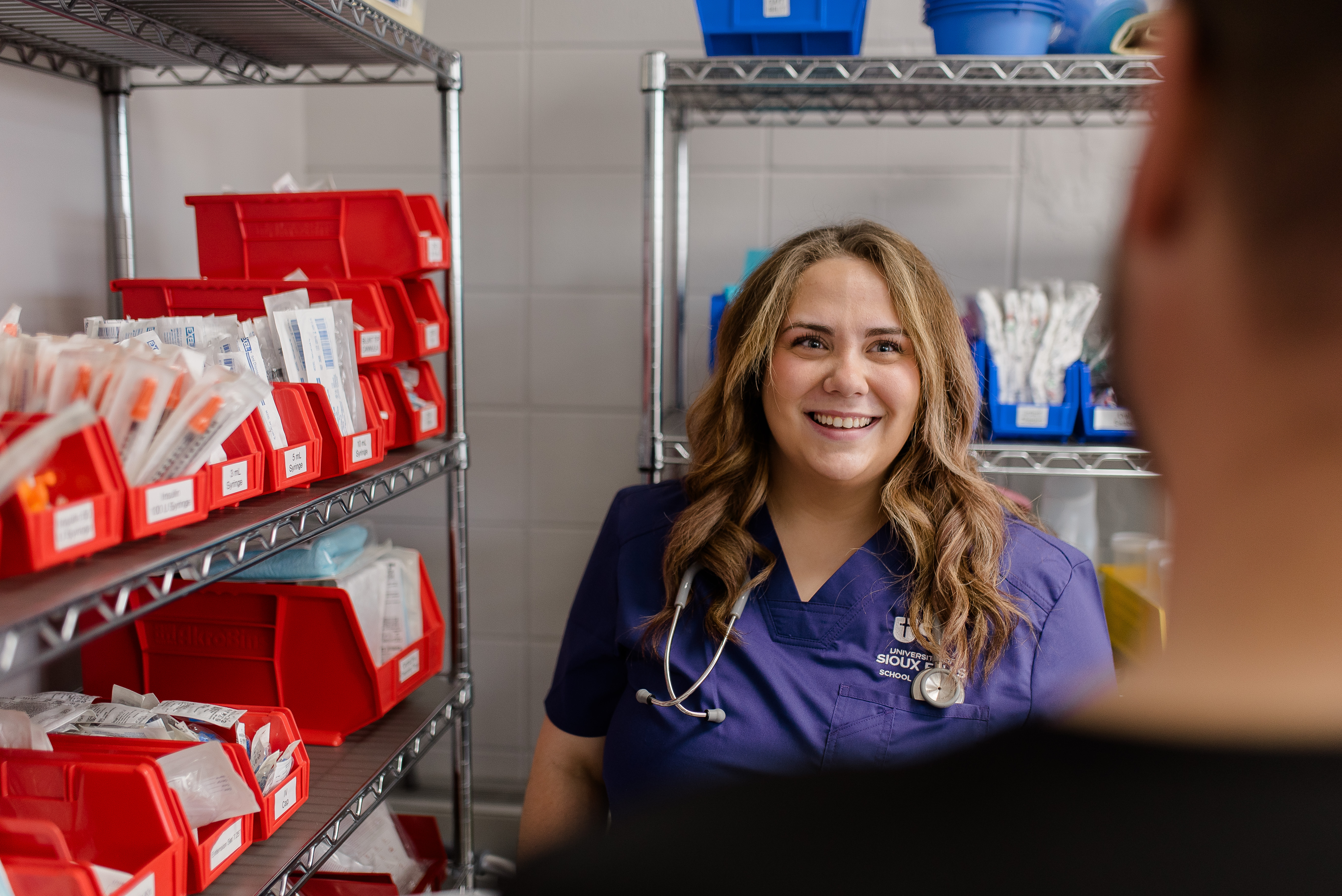 A smiling healthcare worker in a blue uniform with a stethoscope around her neck stands in a medical supply room. She is facing a person whose back is to the camera. Shelves filled with various medical supplies and equipment surround her.